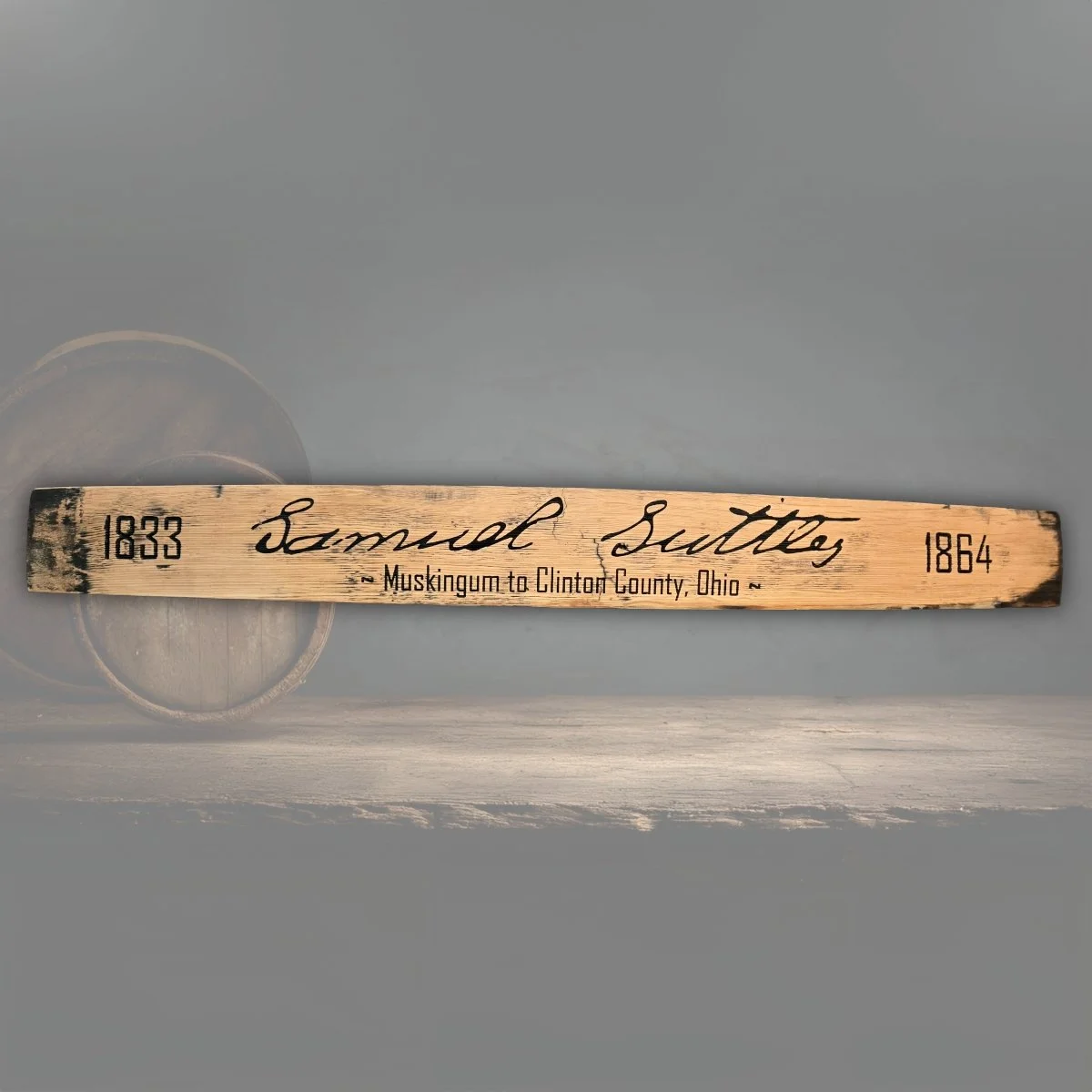 A wooden marker sign with the name Samuel Suttles, showing the years 1833 and 1864, and the text 'Muskingum to Clinton County, Ohio', placed on a wooden surface with a dark background.