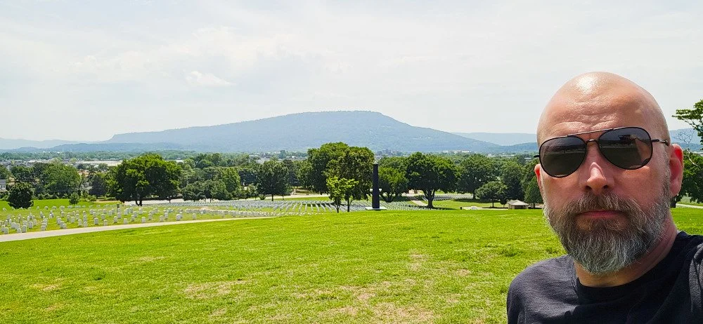 A man with a shaved head, beard, wearing sunglasses and a dark shirt, taking a selfie on a grassy hilltop with a world war II cemetery, trees, and a mountain in the background.