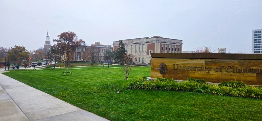 University of Cincinnati campus with a green lawn, trees, historic buildings, and a sign reading 'University of Cincinnati' in the foreground during a cloudy day.