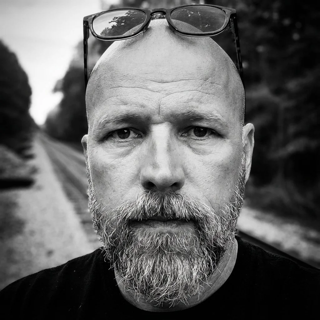 Close-up black and white photo of a bald man with a full beard, wearing glasses on his head, standing outdoors on a railroad track surrounded by trees.