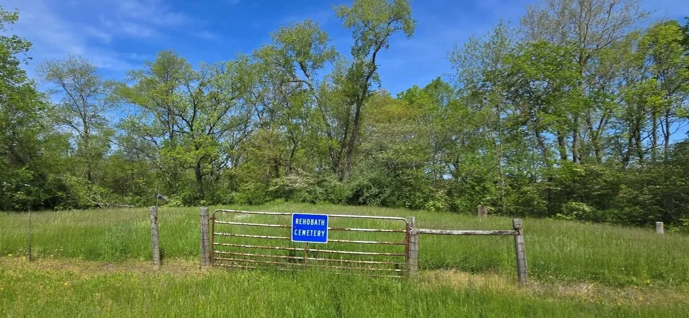 A rusty gate with a blue sign reading 'Rehoboth Cemetery' in front of a grassy field with trees and a blue sky.