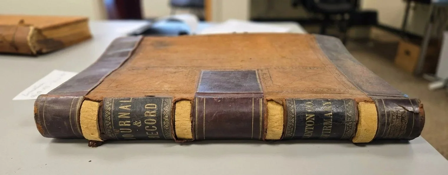 An old, worn leather-bound book with frayed edges and damaged spine, titled 'Journal & Record,' placed on a table with other books and objects in the background.