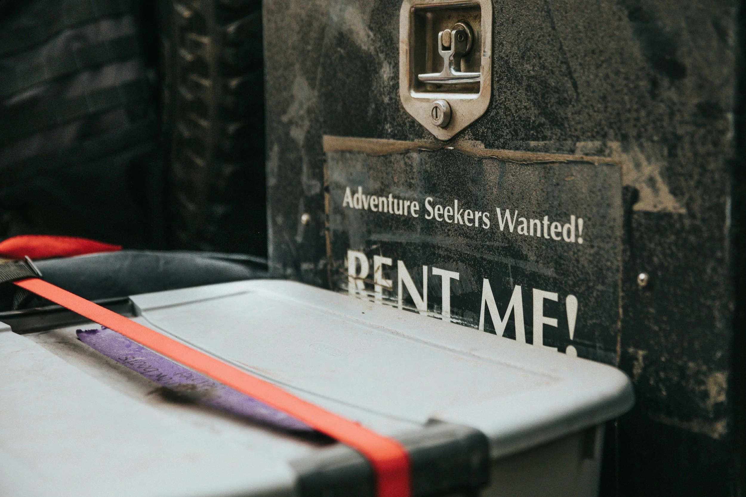 A rental mailbox with a sticker that says "Adventure Seekers Wanted! RENT ME!" in white letters on a black background. There is a plastic storage container with an orange strap in front of it.