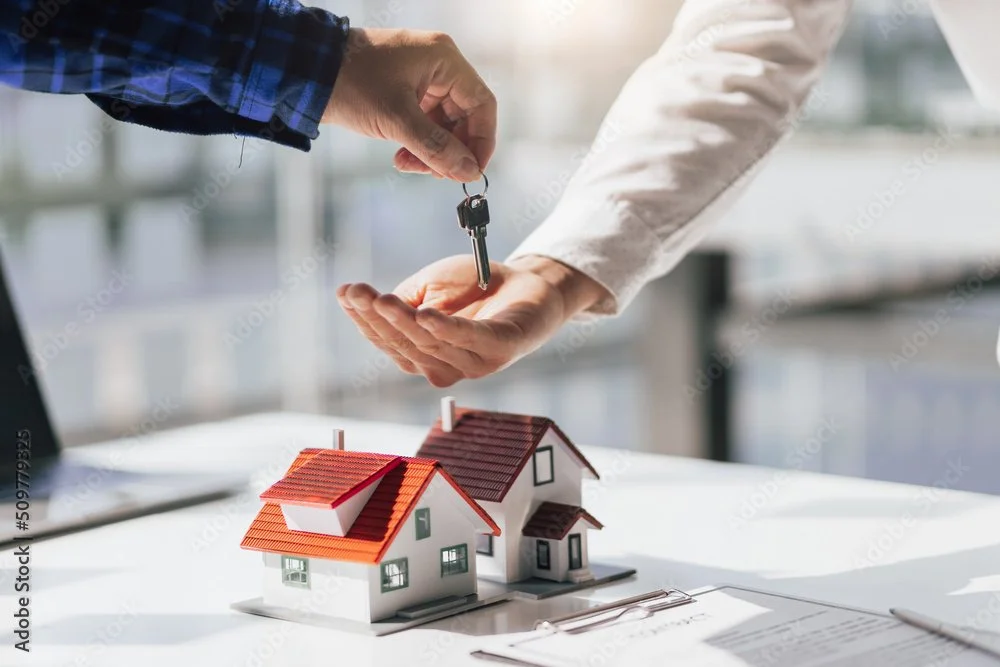 A person is handing over a key to another person over a table with two miniature house models, symbolizing real estate or a property transaction.