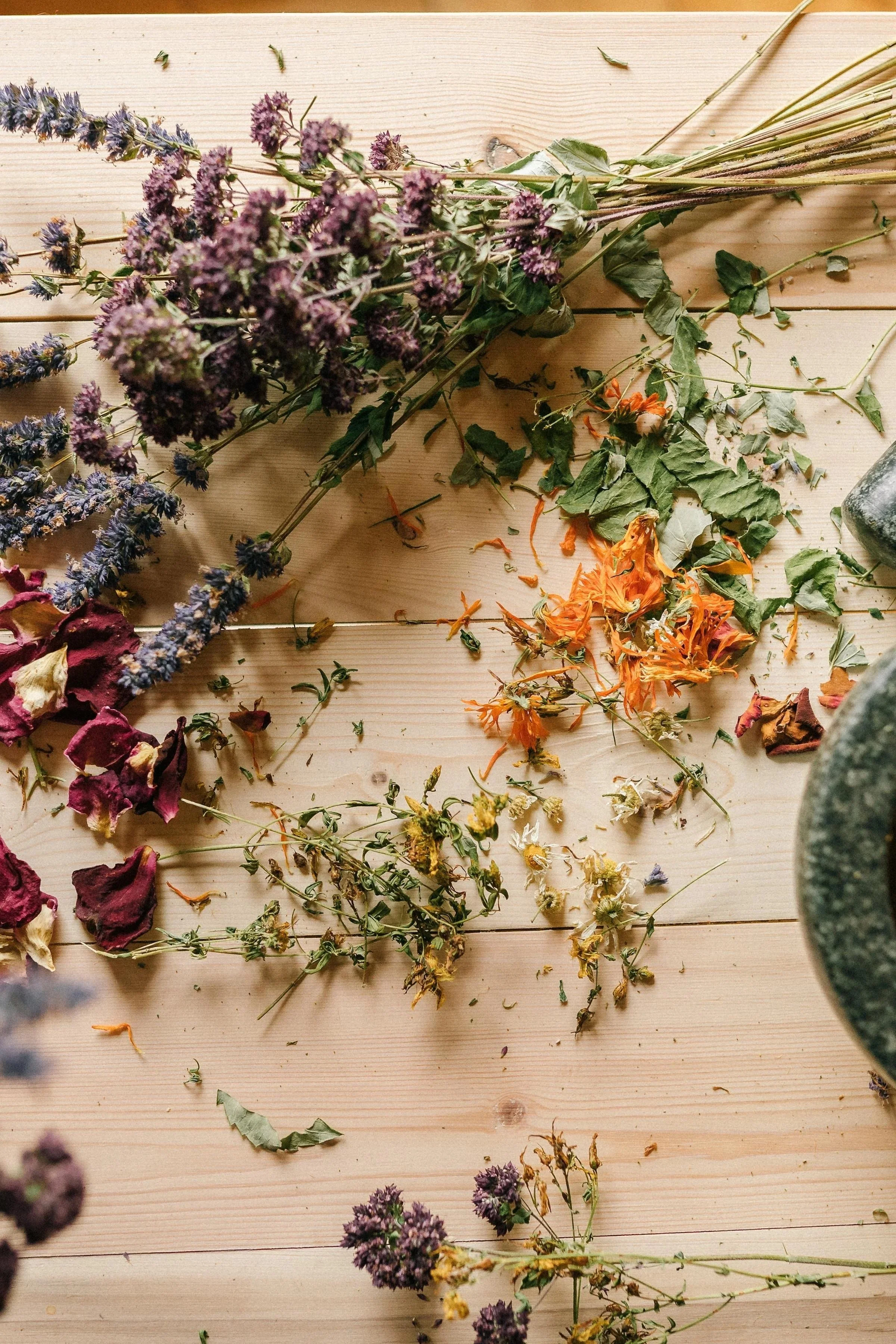 Dried flowers and plant stems scattered on a light wooden surface, including purple lavender, orange marigolds, pink roses, and green leaves.