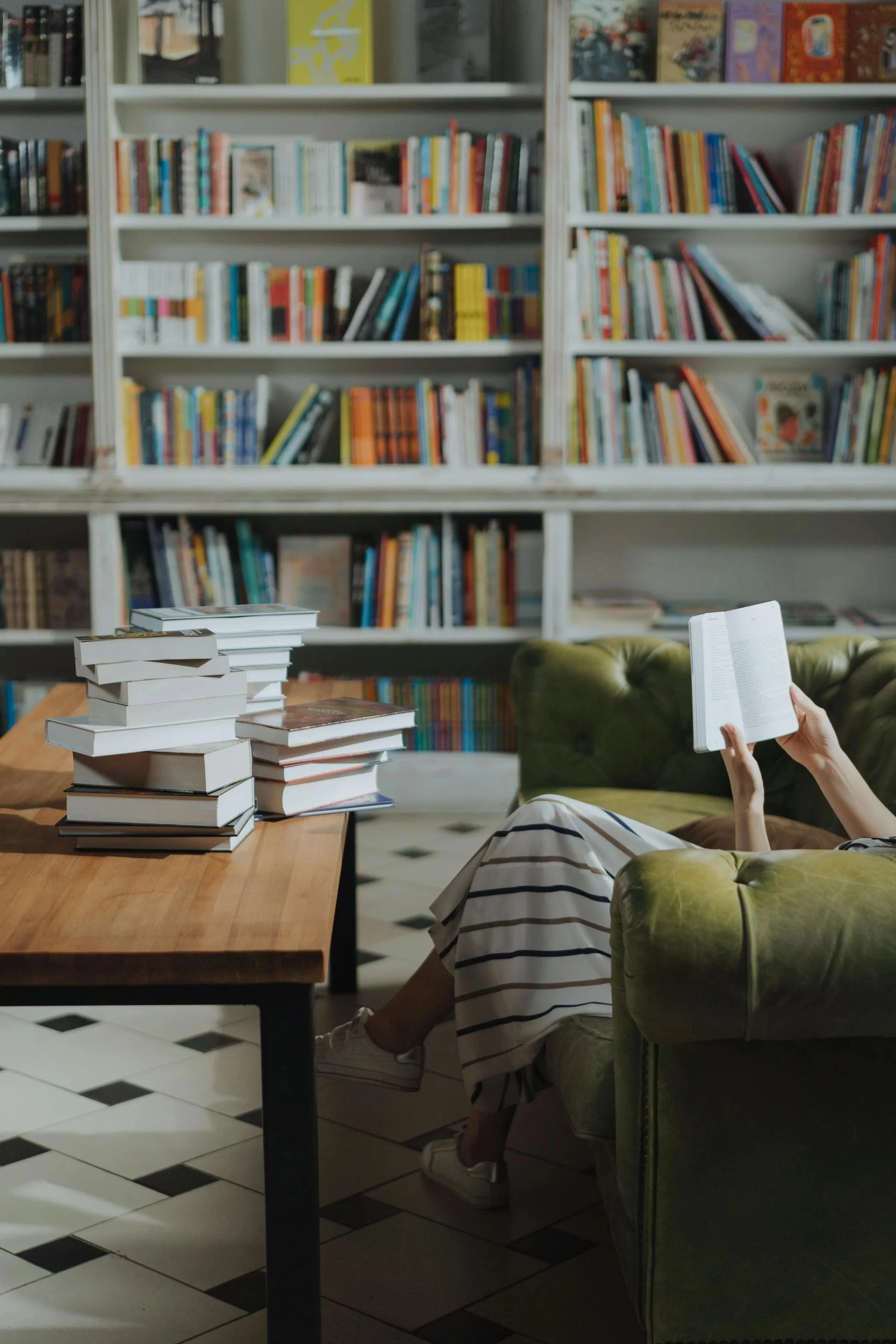Person sitting on a green vintage sofa reading a book in a library, with a table beside them piled with books, and bookshelves filled with colorful books in the background.