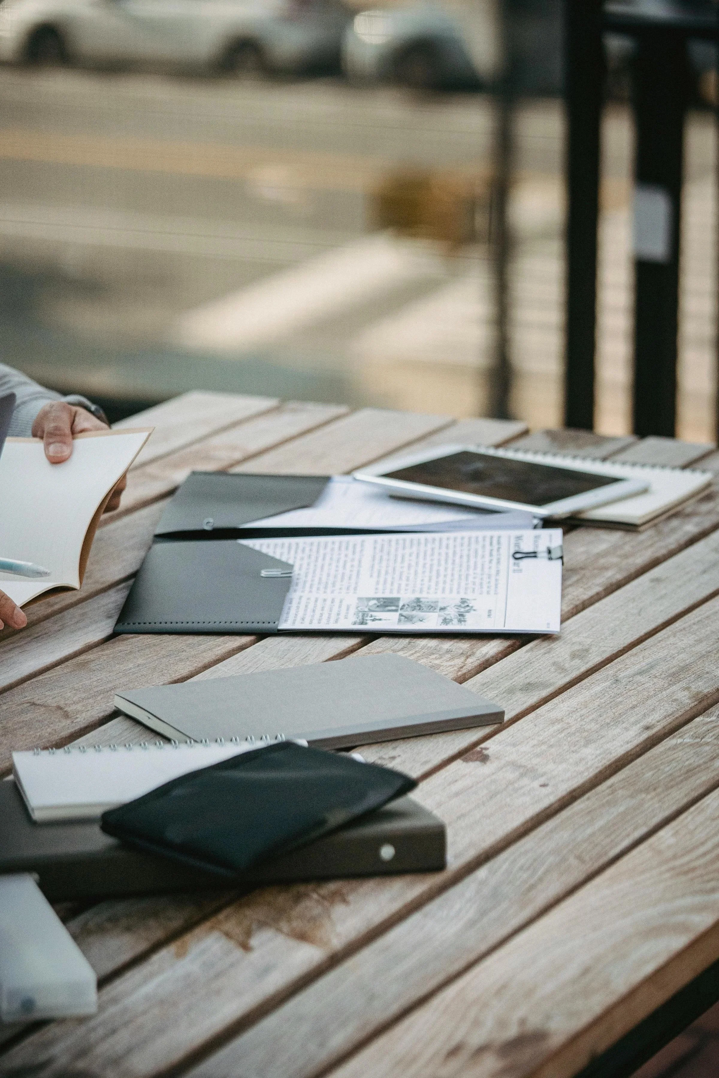 A wooden table with notebooks, papers, and a tablet placed on it, outdoors, with some blurred objects in the background.