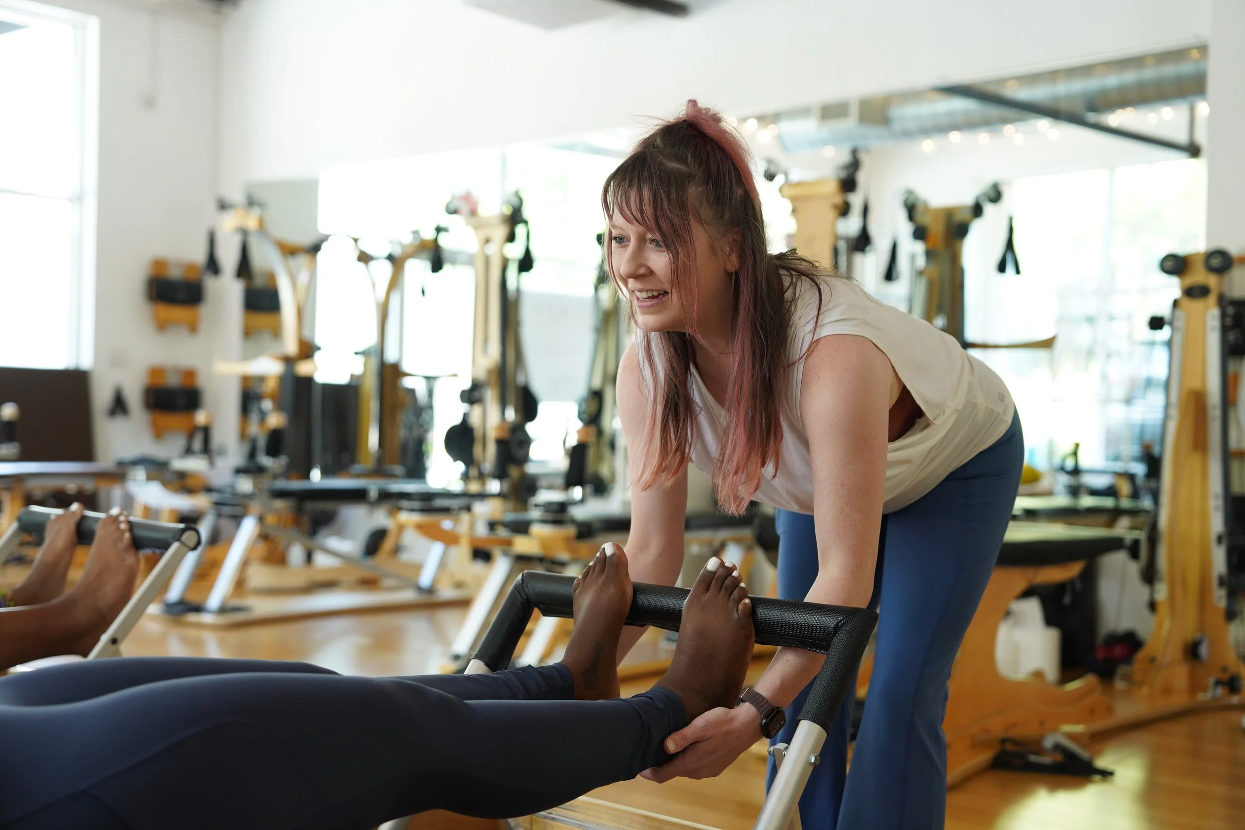 A woman smiling and helping someone during a workout in a gym, with exercise equipment in the background.