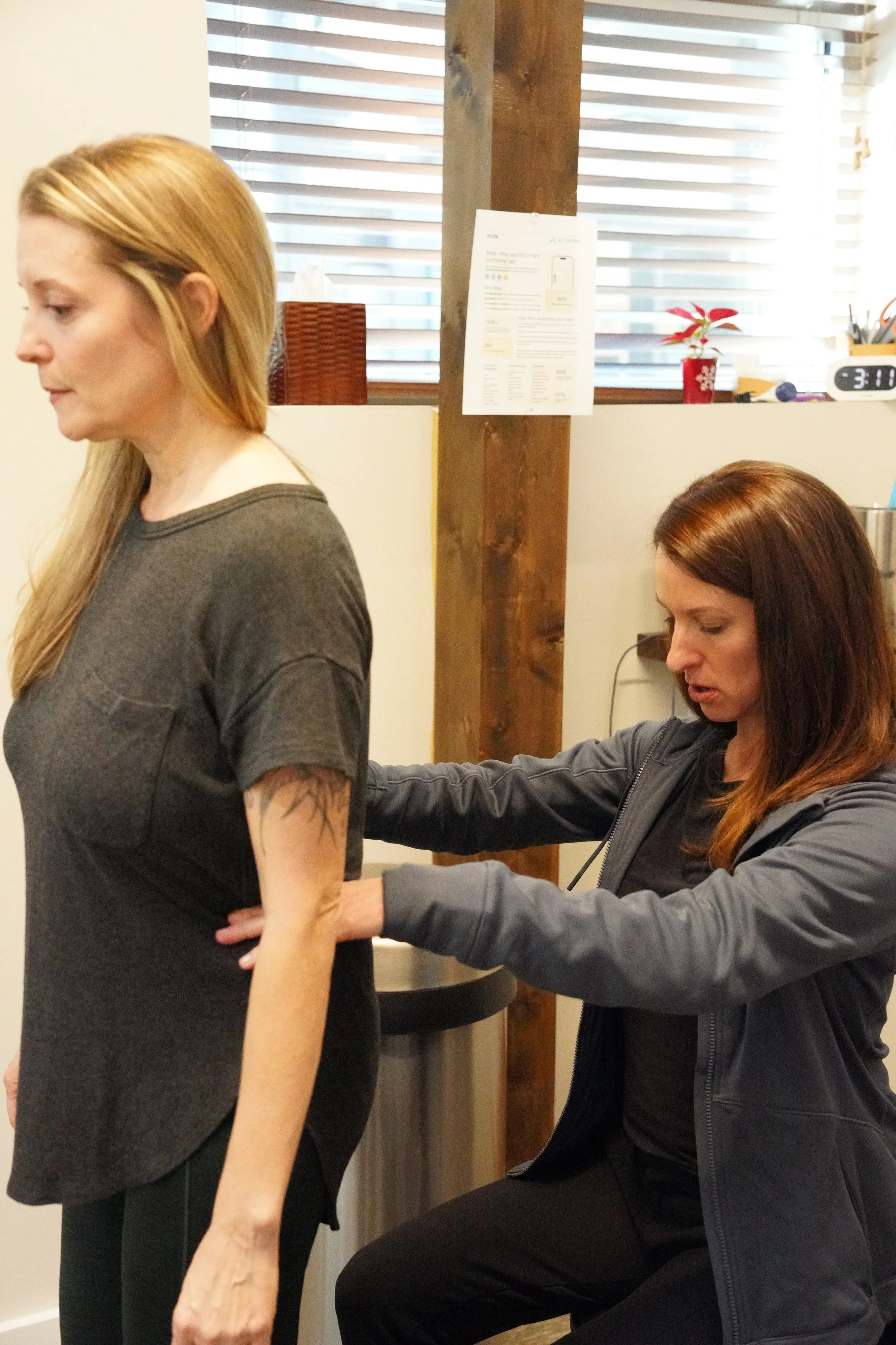 A woman sitting down and feeling the back of another woman standing in front of her in a clinical setting.