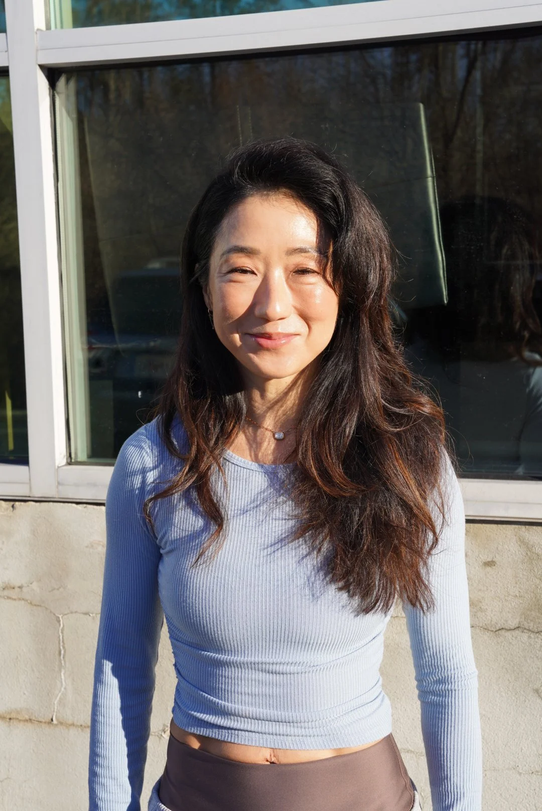 A woman smiling outdoors in front of a window, wearing a light blue long-sleeve top and a necklace.