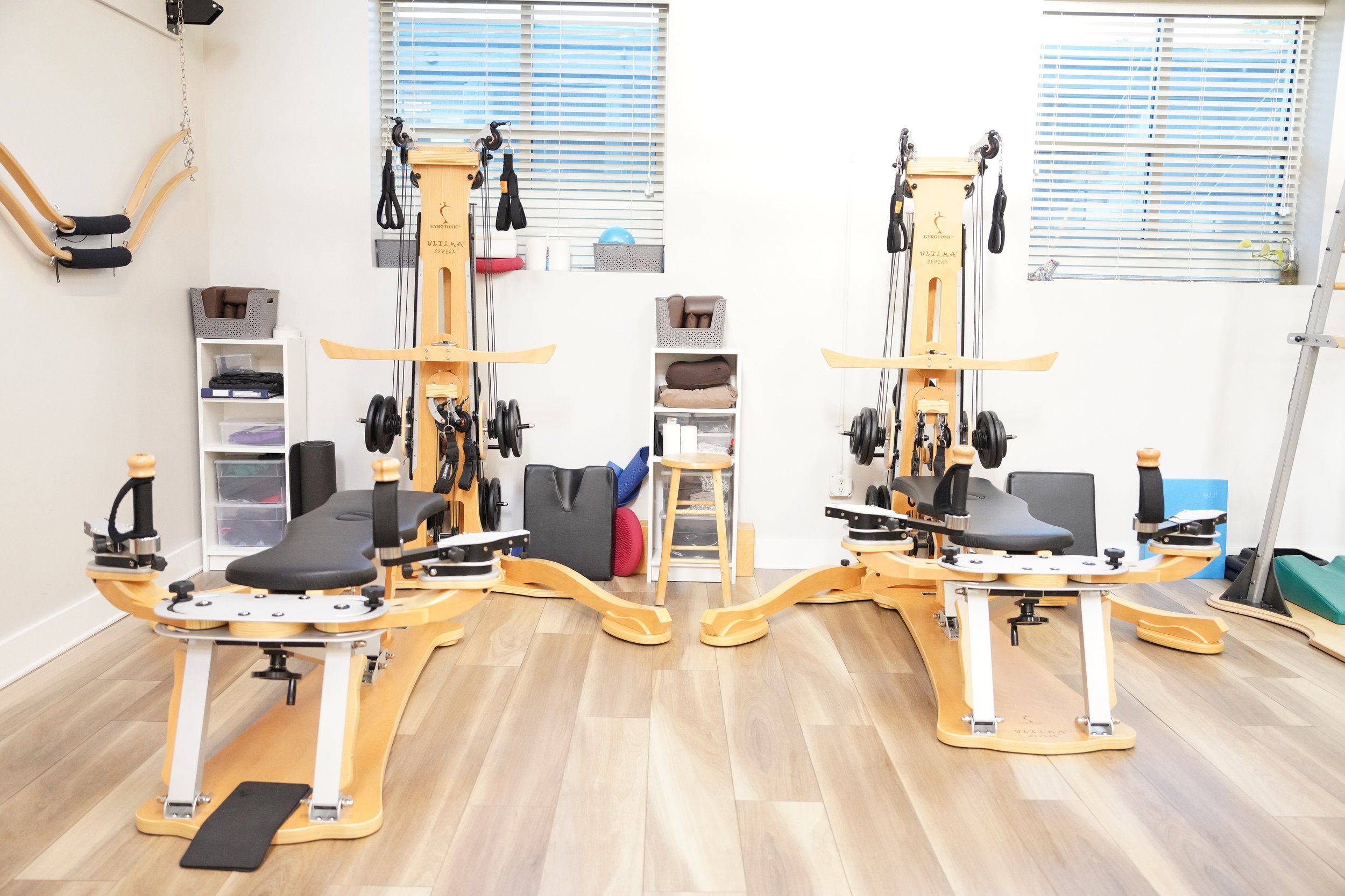 A physical therapy or rehabilitation room with two specialized wooden exercise machines, foam rollers, and various fitness equipment, with blinds covering windows in the background.
