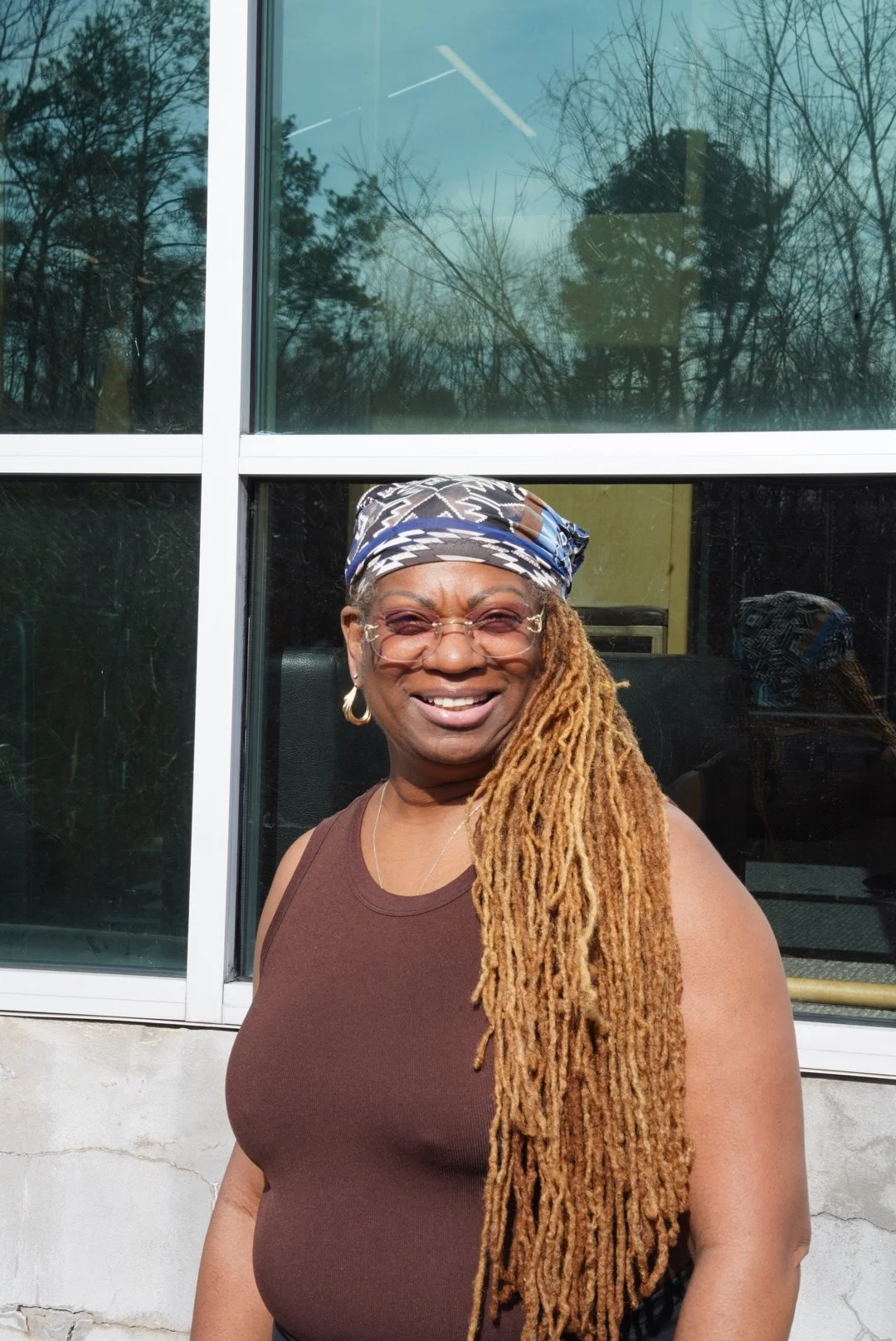 A smiling woman with long blonde dreadlocks, wearing glasses, hoop earrings, a patterned headscarf, and a brown sleeveless top, standing outside near a building with large glass windows reflecting trees.