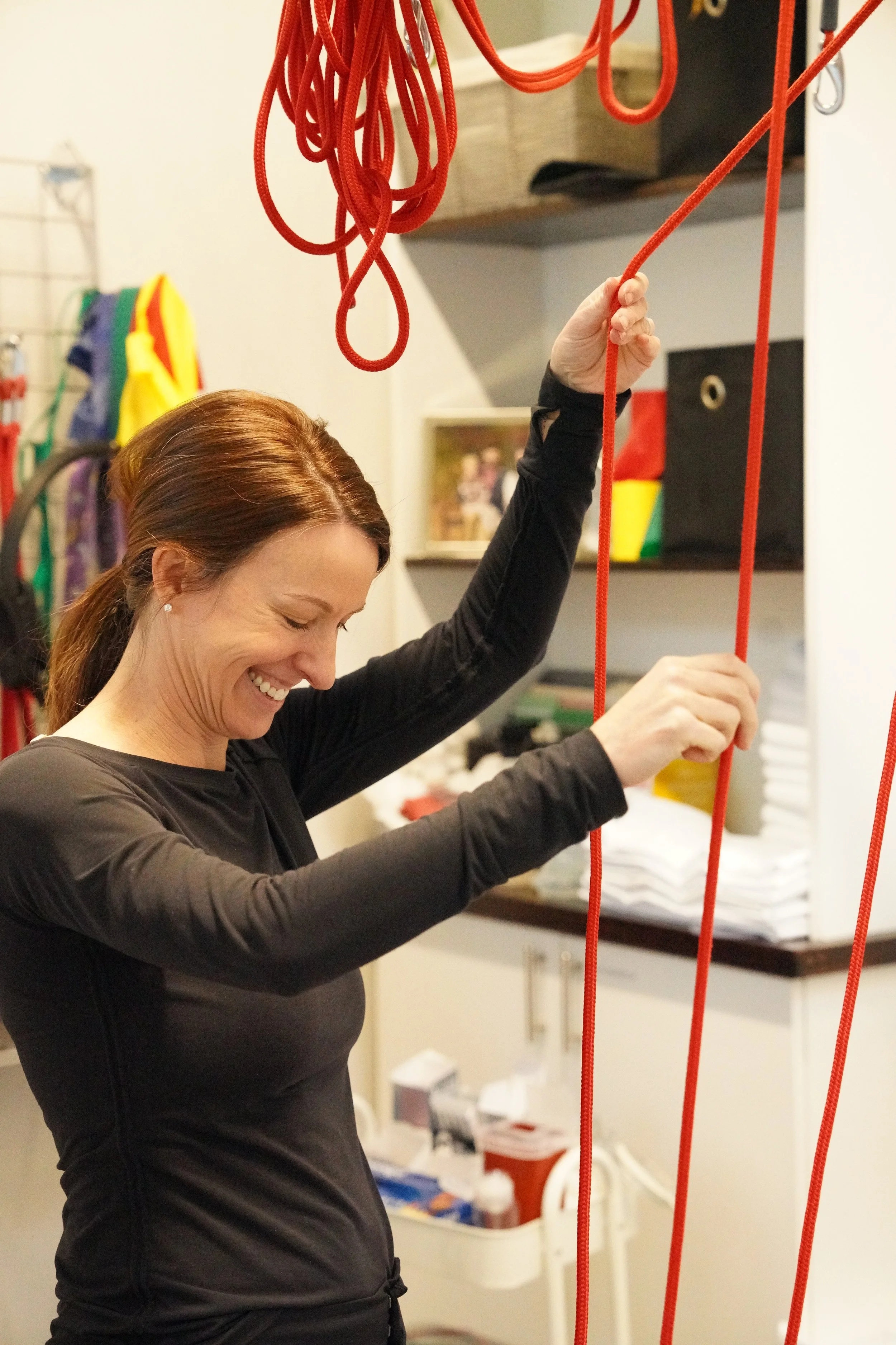 A woman with brown hair, wearing a black athletic shirt, smiling while holding an orange climbing rope at an indoor climbing gym.