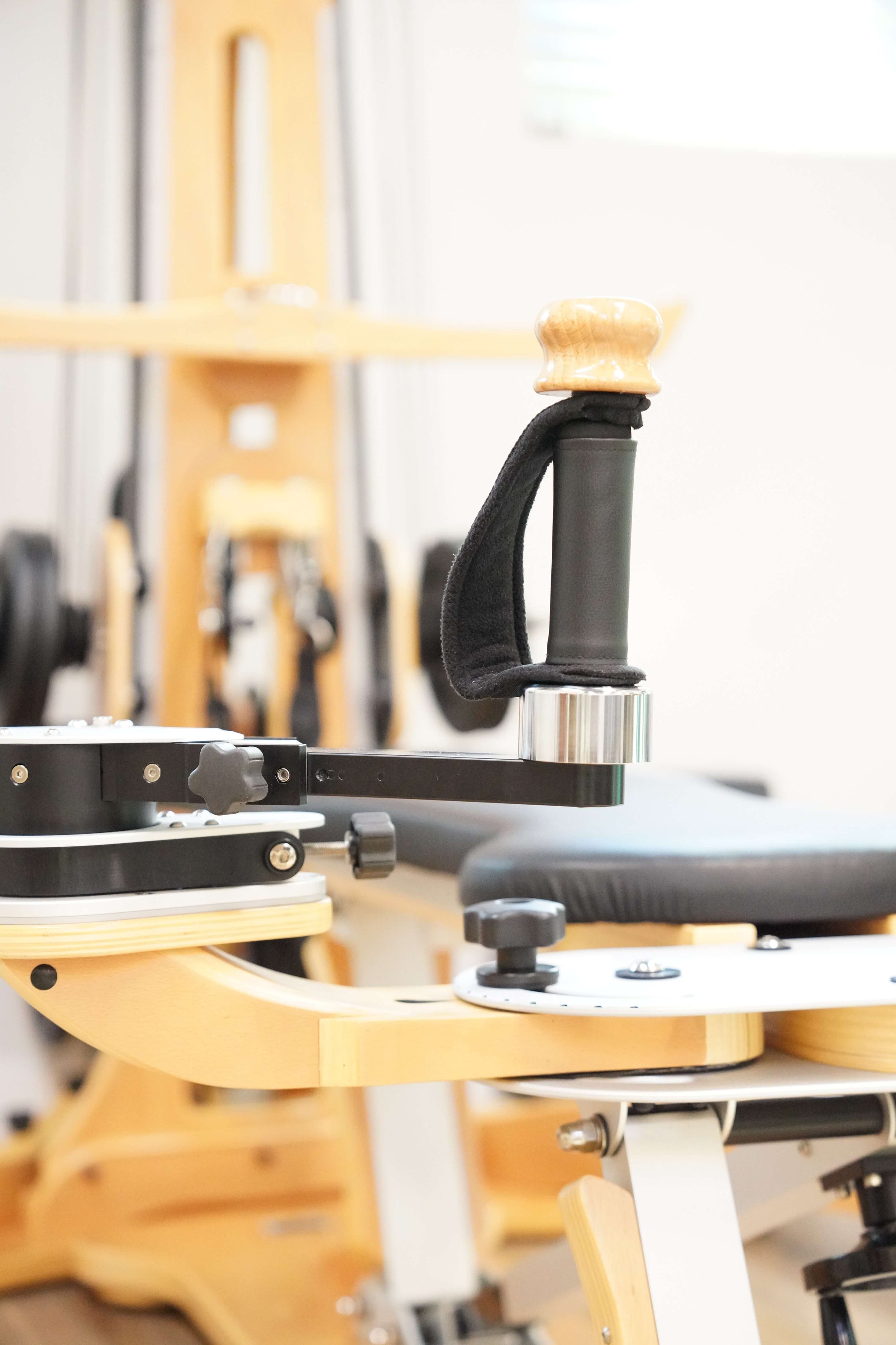 Close-up of a specialized wheelchair or medical equipment with a wooden handlebar and black padding in a room with weightlifting equipment and a chair in the background.
