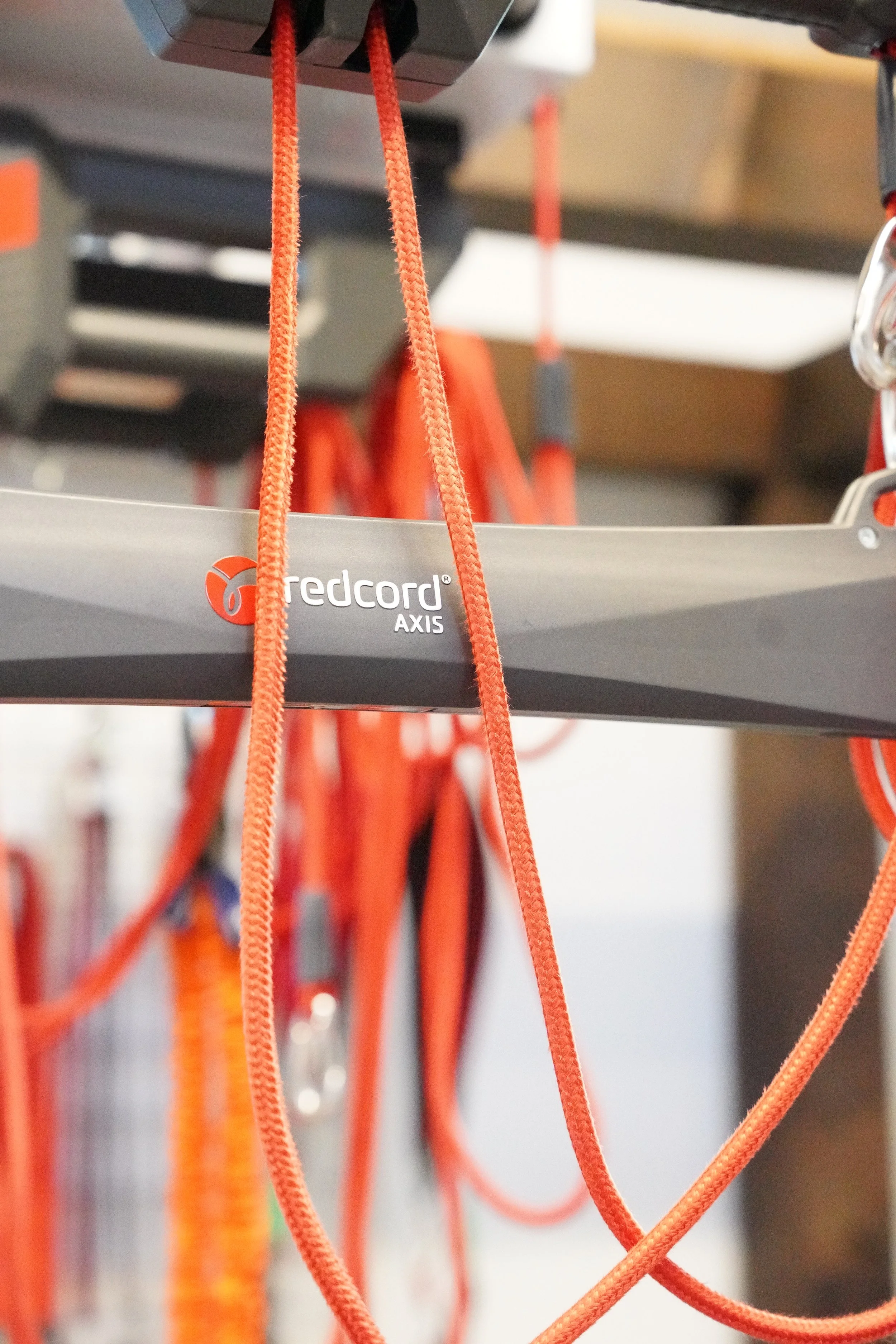 Close-up of orange climbing ropes hanging from a gray 'Redcord AXIS' equipment station in a gym or training facility.