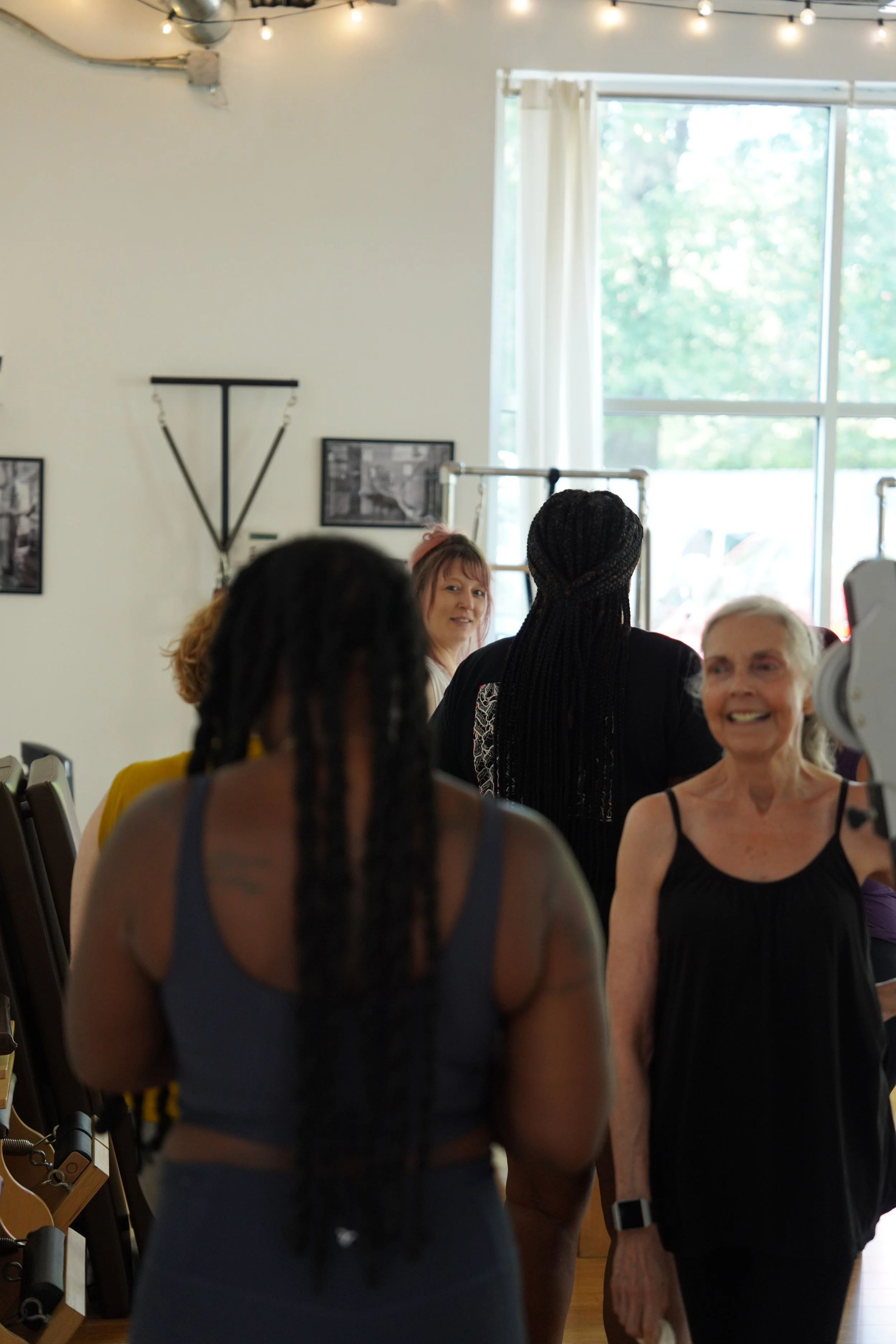 A group of women gather in a bright room with large window, white curtains, and string lights on the ceiling, engaging in social interaction.