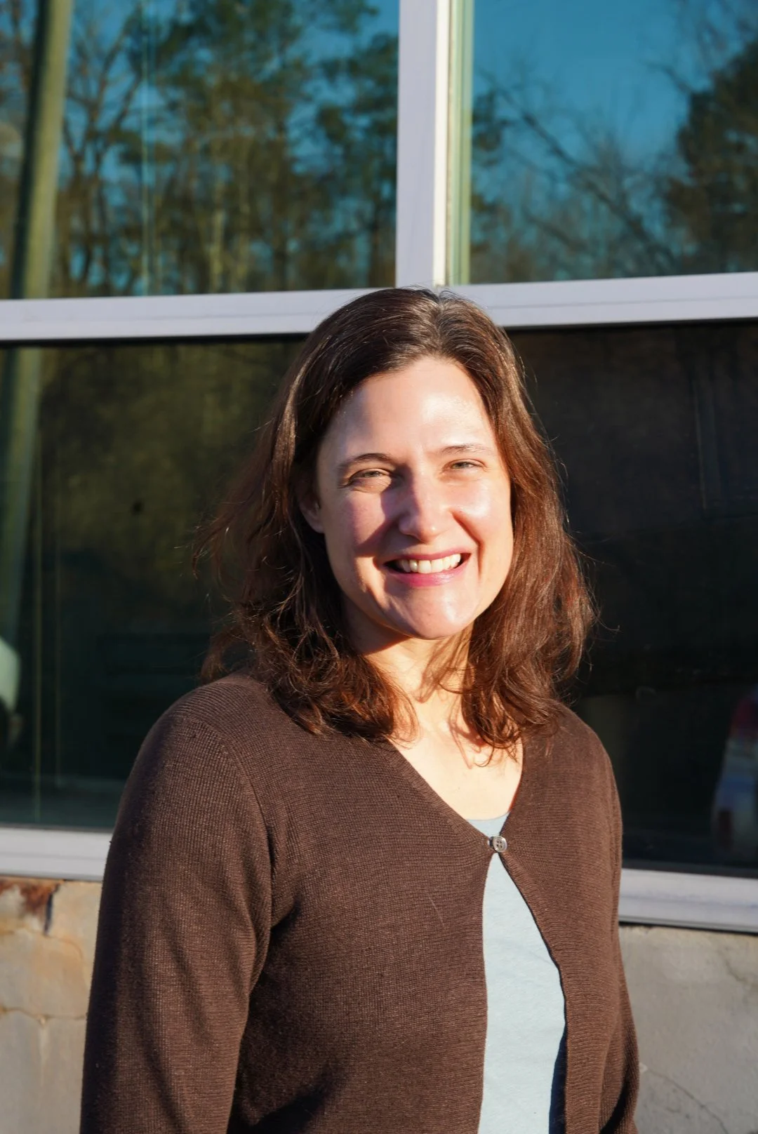 A smiling woman with shoulder-length brown hair standing outdoors near a building with large windows, reflecting trees and the sky in the background, sunlight illuminating her face.