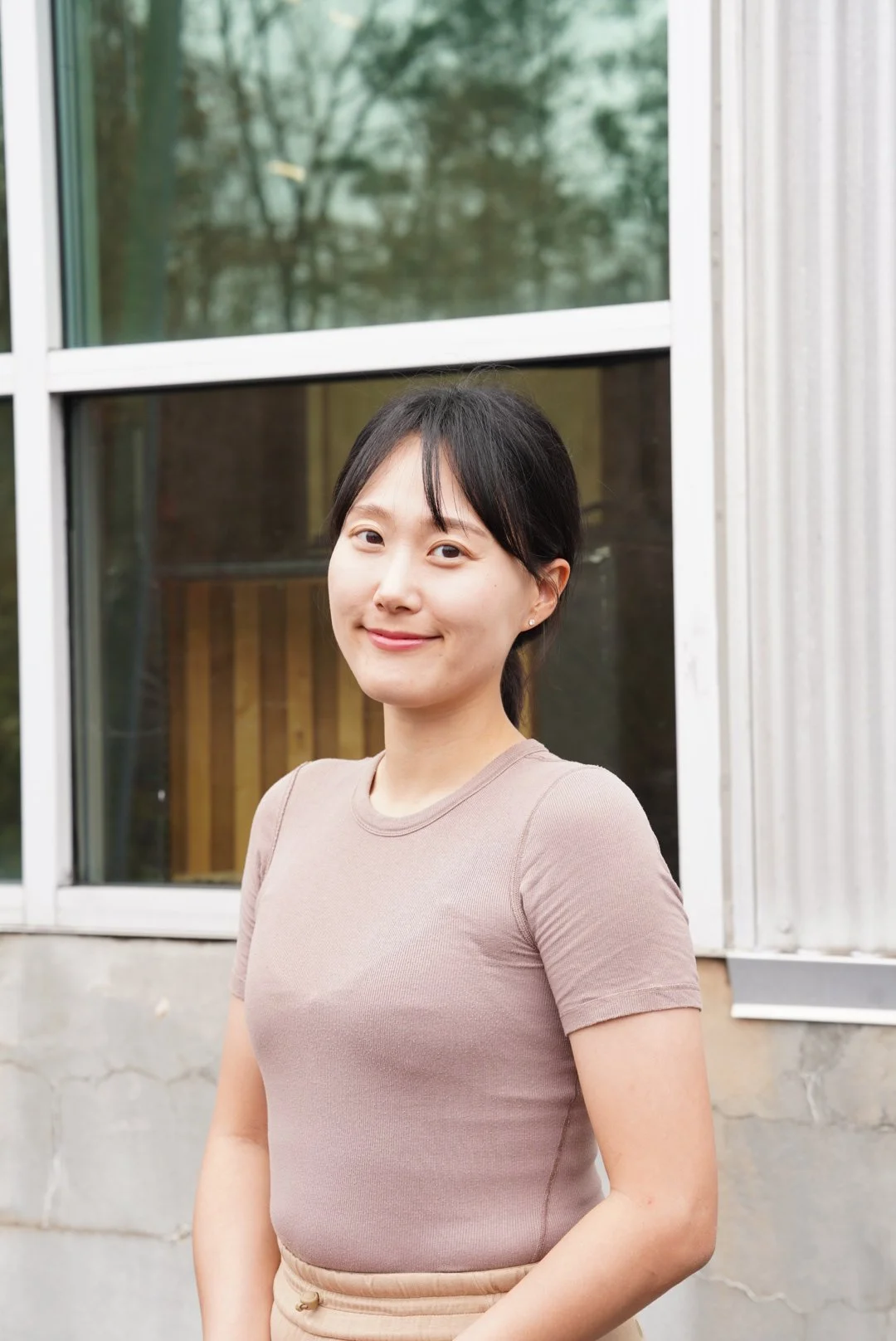 A woman standing outdoors near a building with large window panes, smiling at the camera.