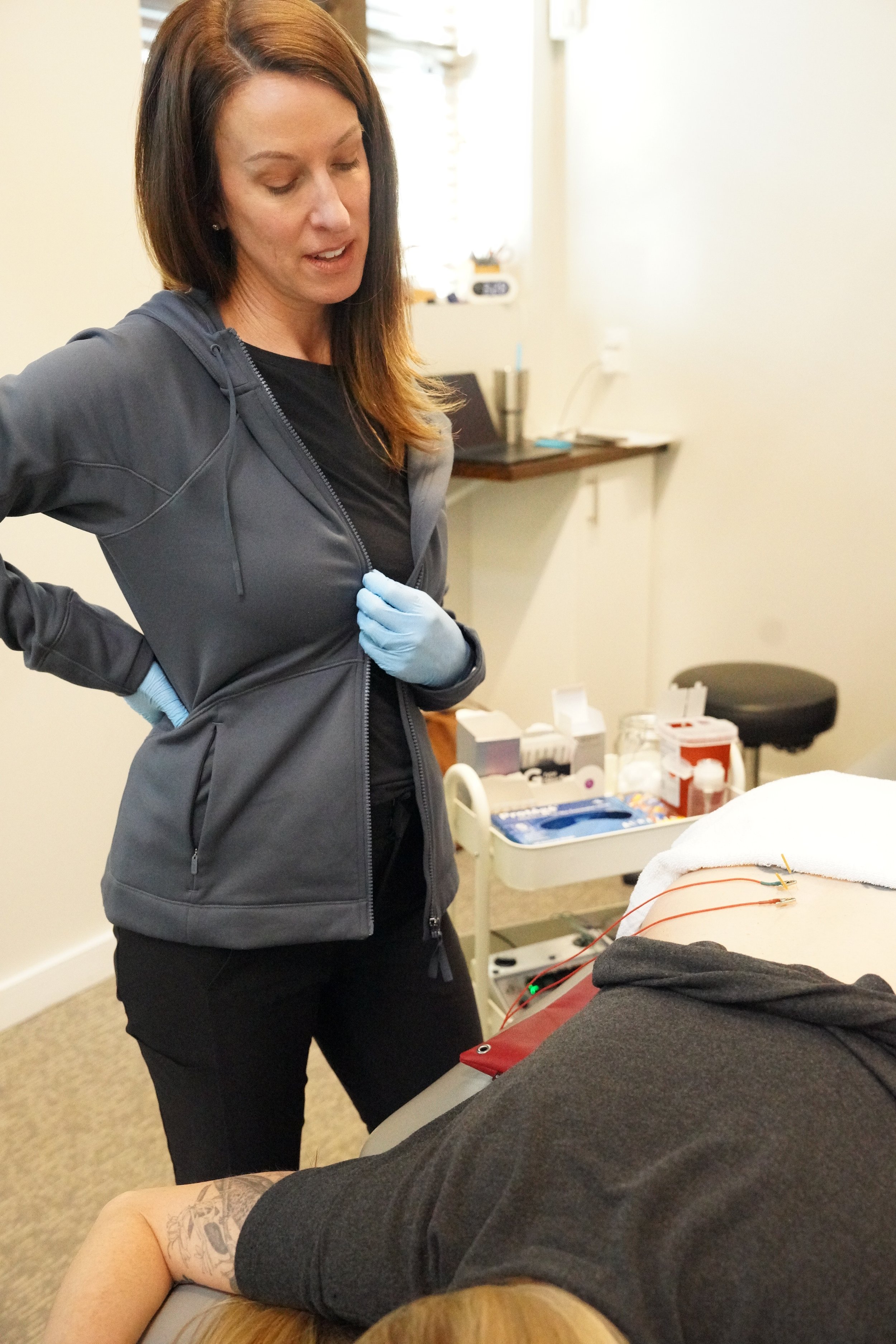 A woman in a gray hoodie and gloves performing a medical procedure on a patient's arm, which is connected to electrodes. The setting appears to be a healthcare or clinical environment with medical supplies on a tray and a whiteboard in the background.