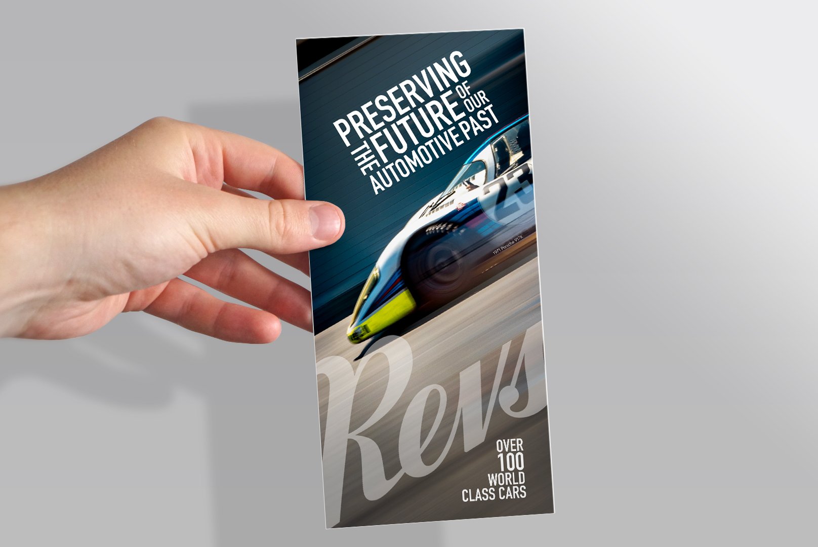 A hand holding a booklet or brochure titled 'Preserving the Future of our Automotive Past,' with a picture of a high-speed train on the cover and text mentioning 'over 100 world class cars' in the background.