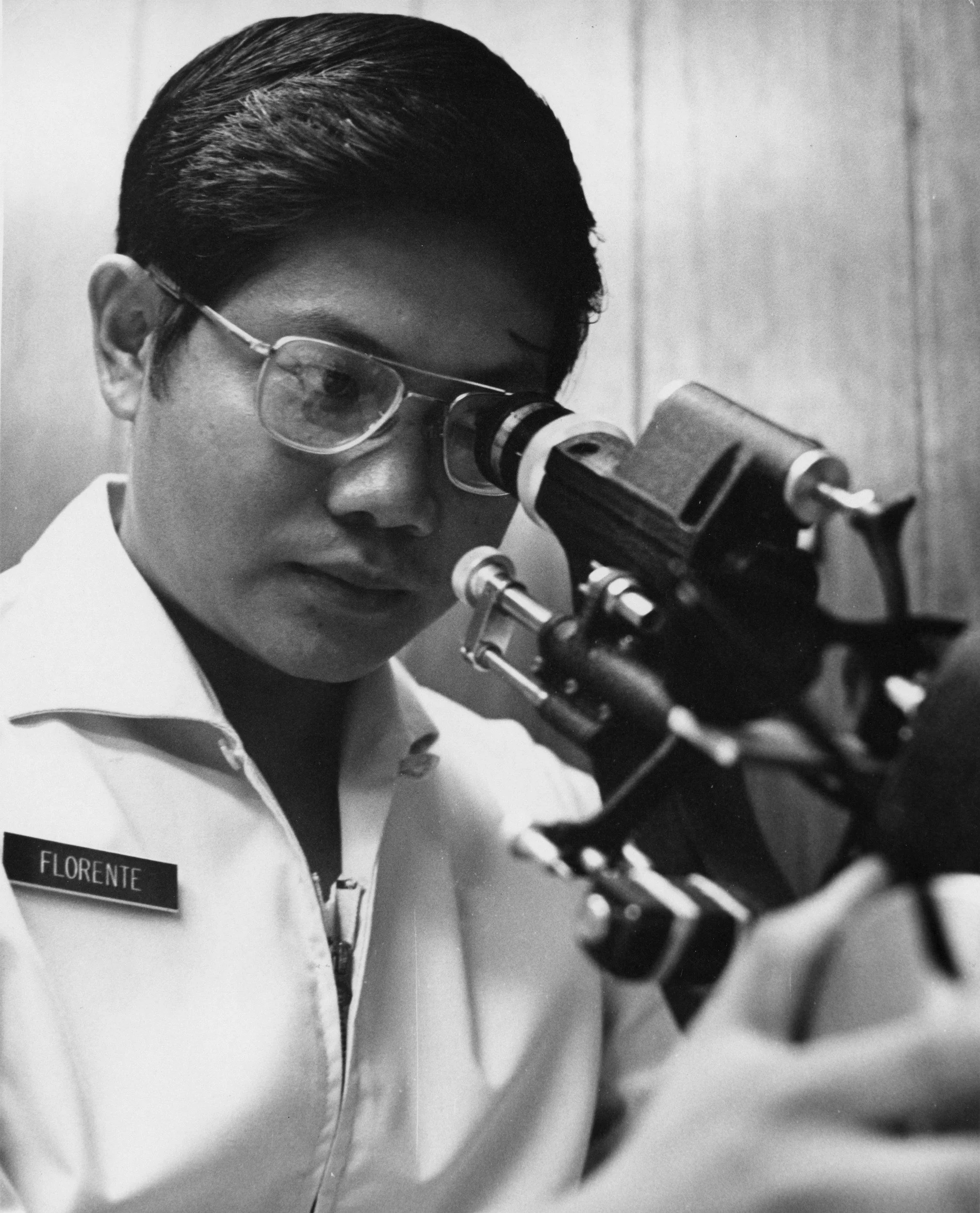 Black and white photo of a woman looking through a microscope, wearing glasses, a white lab coat with a name tag that says 'FLORENTE'.