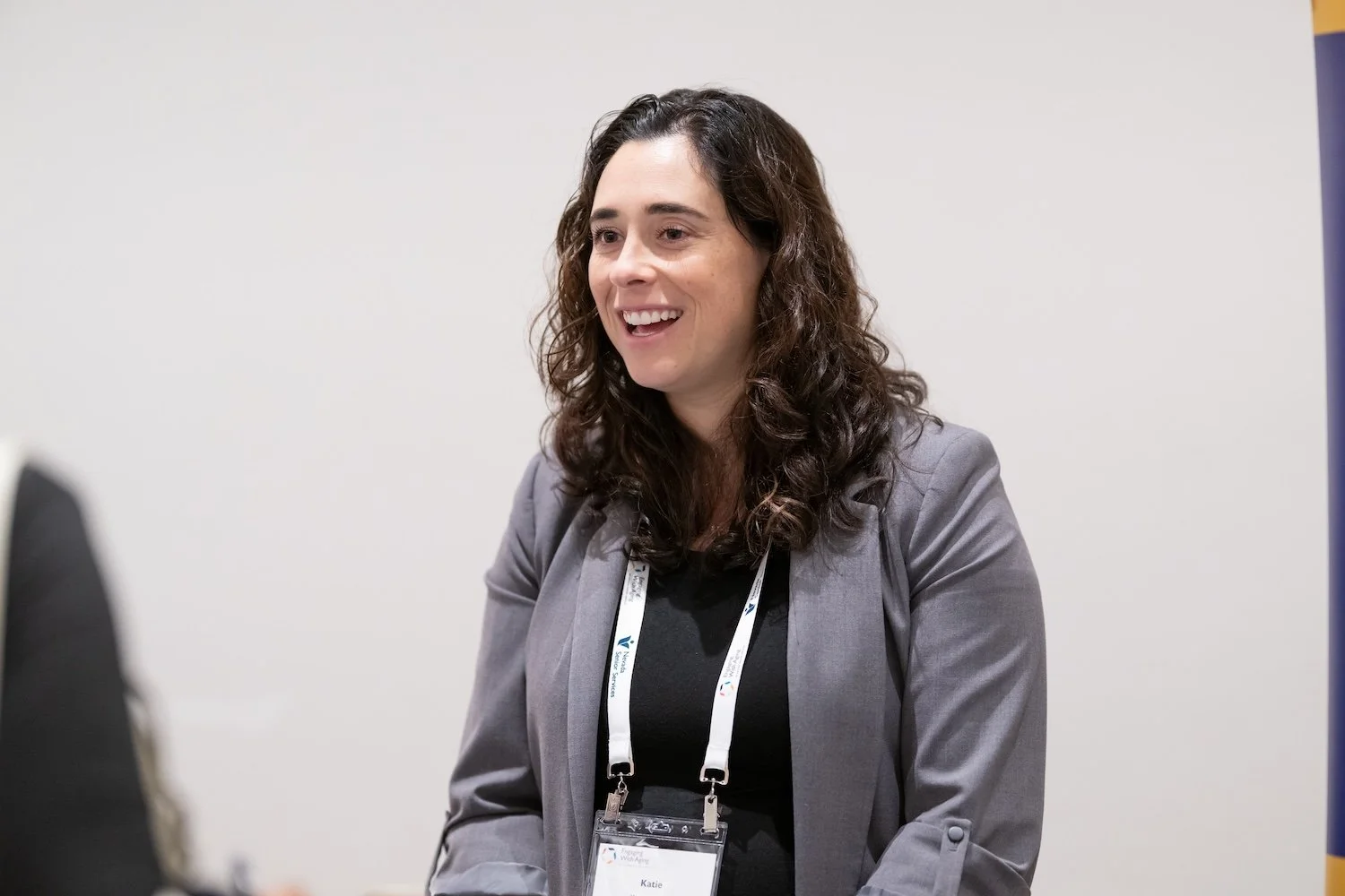 A woman in a black leather jacket smiling as she hands a badge or pass to a woman with long braided hair, wearing a plaid pants and a beige top, at a registration or check-in table with many black drawstring bags and documents. In the background, people are lined up inside a warmly lit indoor space with large windows.