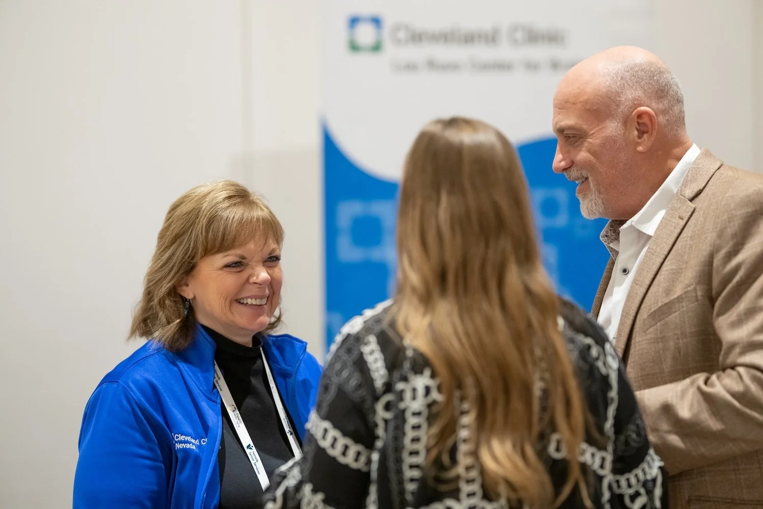 A large conference room filled with people sitting in beige chairs, facing a screen displaying a presentation titled 'Questions?' with contact information for Nevada regional trainers. Four women are standing near the front, one speaking to the audience, and another talking with a woman in front. The room has a wood-paneled ceiling and a plain cream-colored wall.
