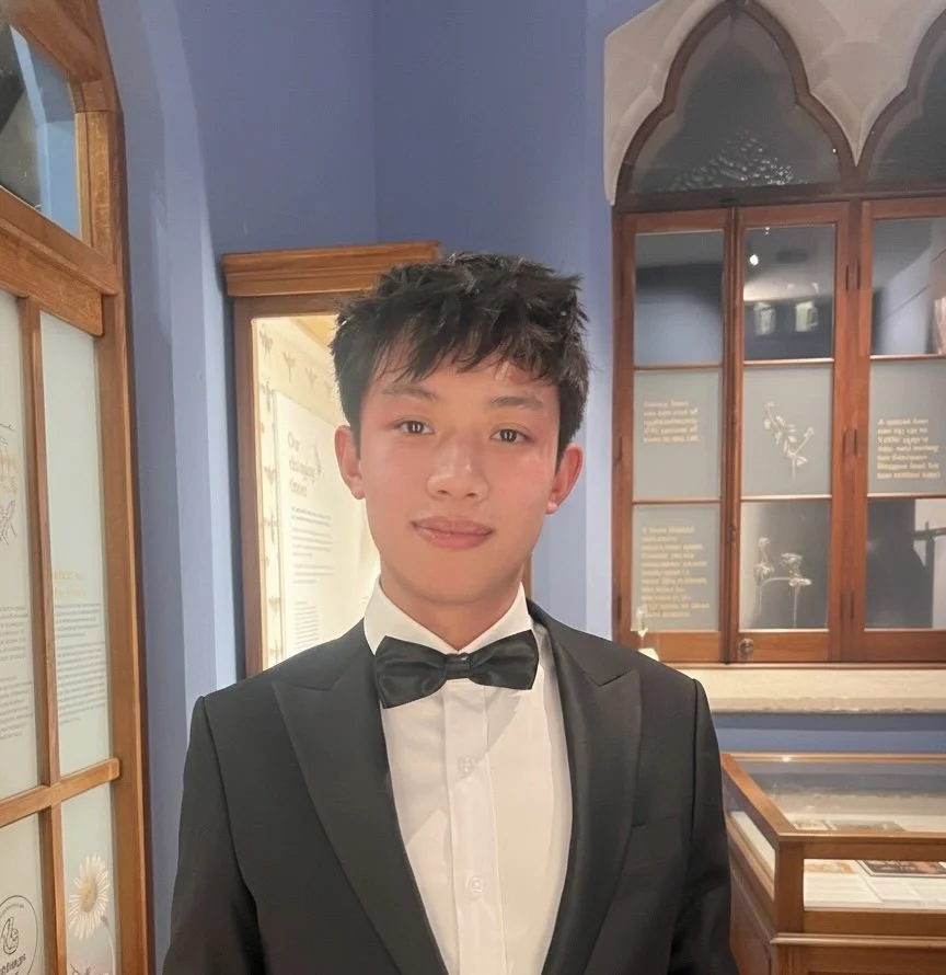Young man in a black tuxedo with a black bow tie, standing in a room with blue walls and wooden framed display cases.
