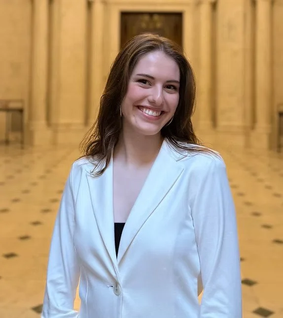Junge Frau mit braunen Haaren und weißem Blazer, lächelnd, vor einem goldenen Theater oder Konzertsaal.