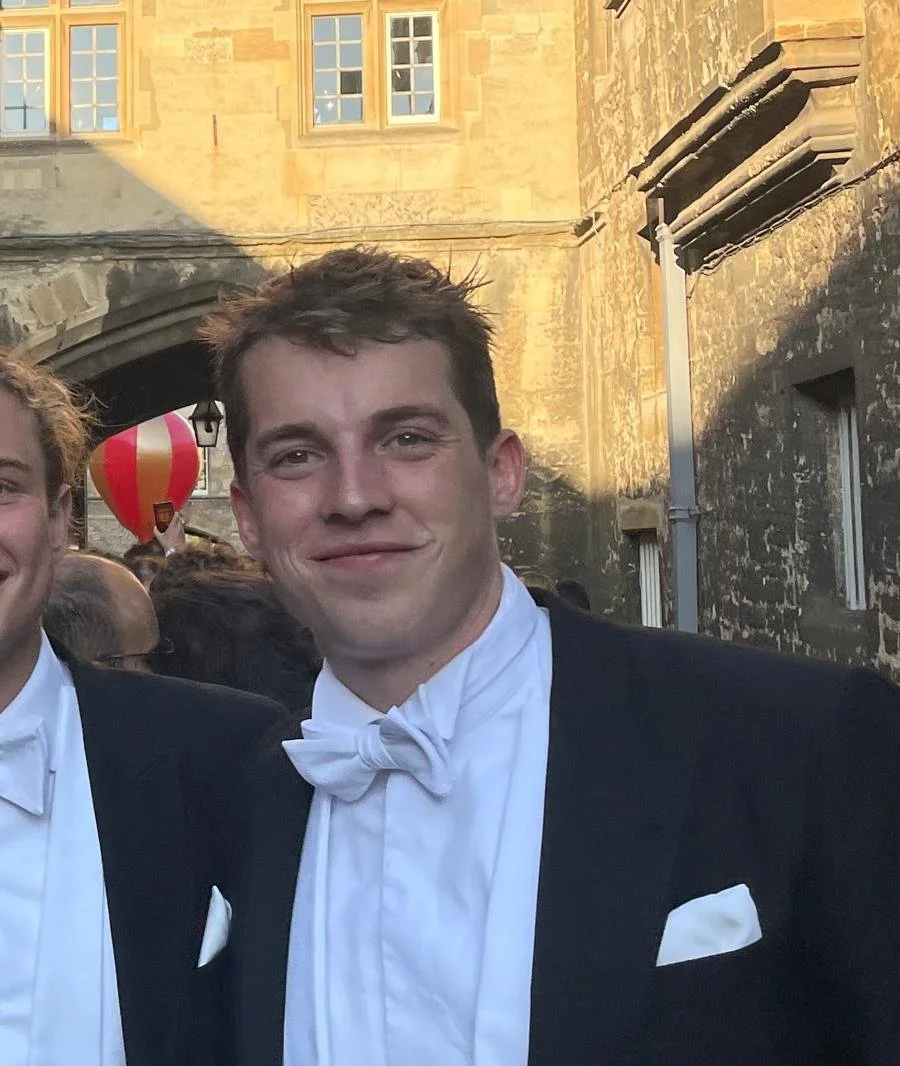 Portrait of a young man in a tuxedo with a white bow tie, smiling outdoors during the evening, with historical stone buildings in the background and a red and yellow hot air balloon in the distance.