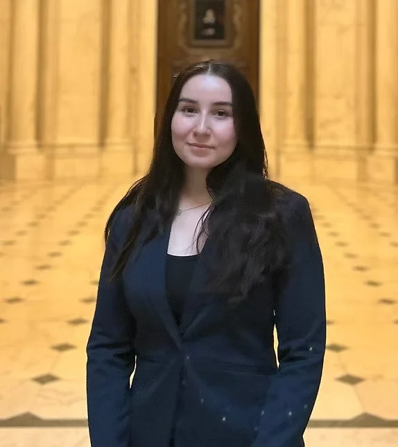 Junge Frau mit dunklen Haaren in einem schwarzen Blazer vor einer Holzvertäfelung im Saal.