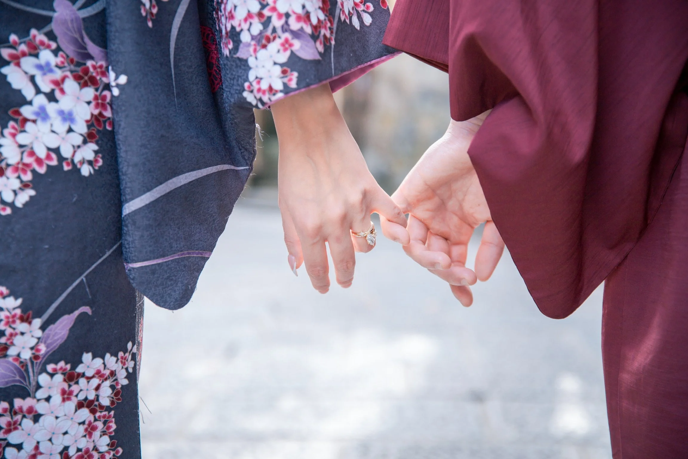A couple holding hands, one wearing a floral kimono and the other in a maroon kimono, with a focus on their interlinked fingers and a ring on the woman's finger.