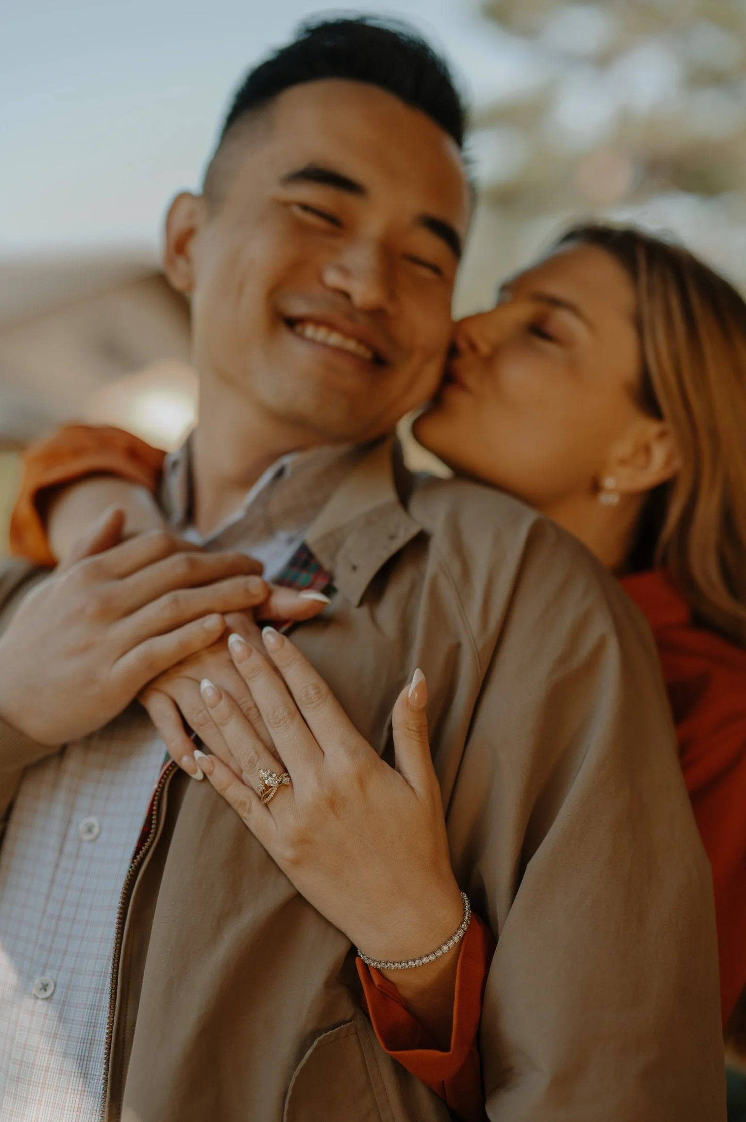 A happy couple sharing a kiss and showing off an engagement ring on the woman's finger.