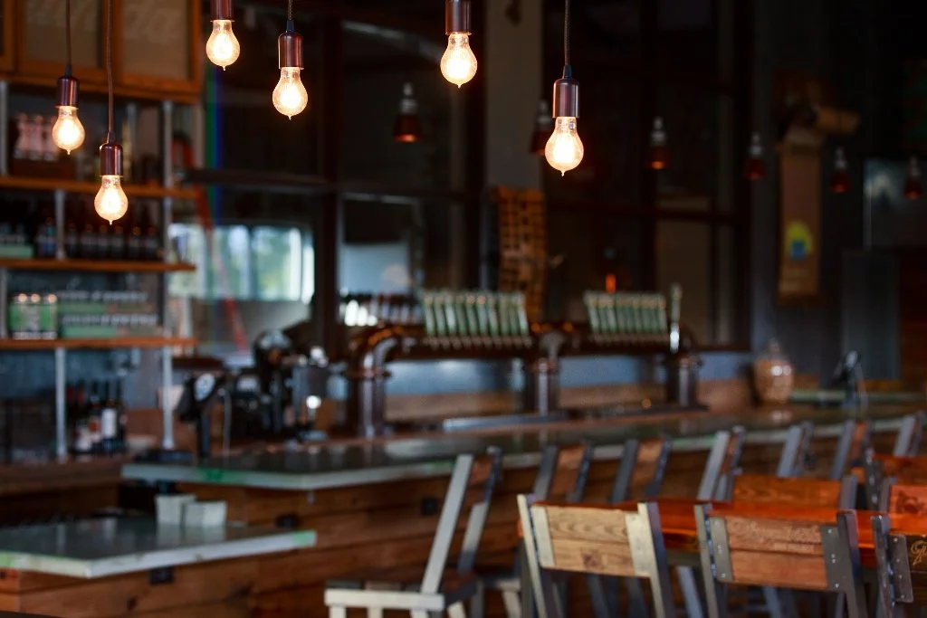 Empty bar with wooden tables and chairs, hanging exposed light bulbs, shelves with bottles and glasses, and a coffee espresso machine in the background.