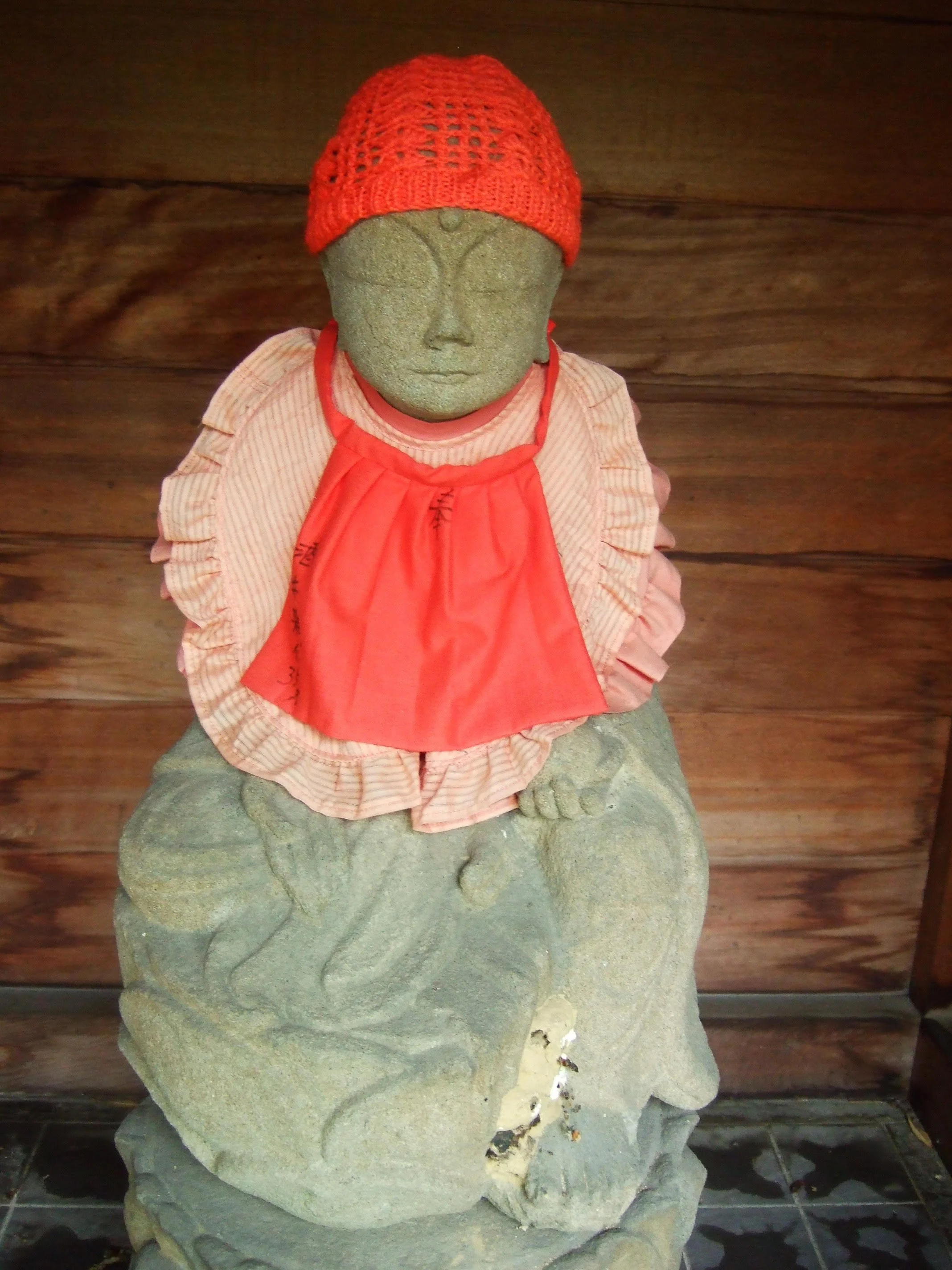 A stone Jizo statue wearing a red knit hat, a pink and white striped shirt, and a red scarf, seated on a carved stone pedestal against a wooden wall background.