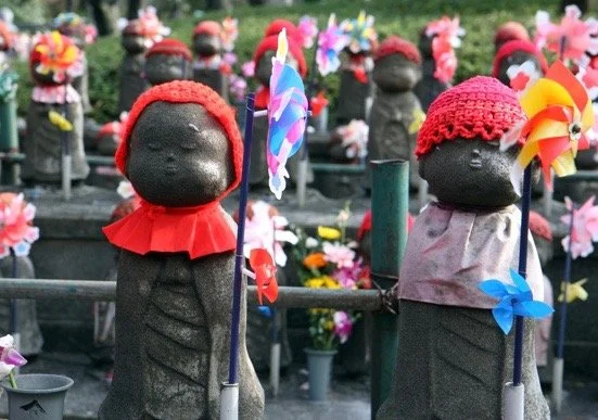 Jizo stone statues dressed as children with knitted hats and colorful pinwheels, surrounded by other similar statues in a garden or memorial setting.