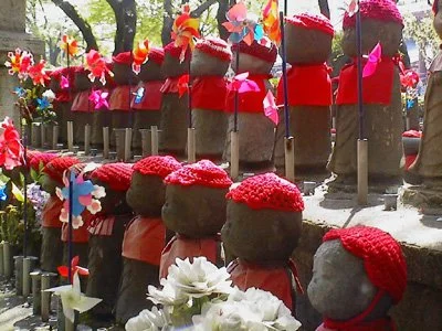 Row of small stone Jizo statues decorated with red hats and colorful windmill toys, surrounded by flowers and trees.