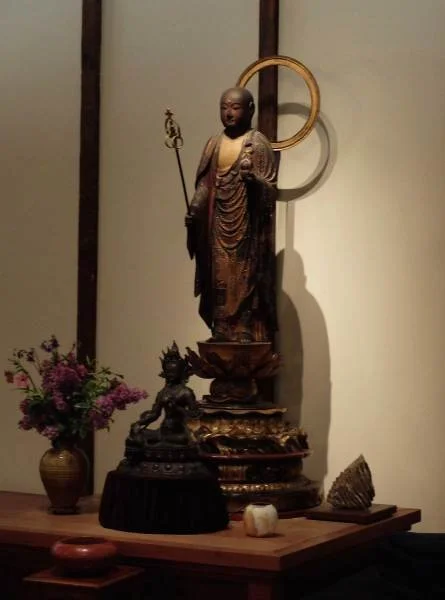 A wooden Buddha statue with a circular halo, holding a staff, on a decorated platform with smaller statues, a flower vase, and a shell, against a plain wall.