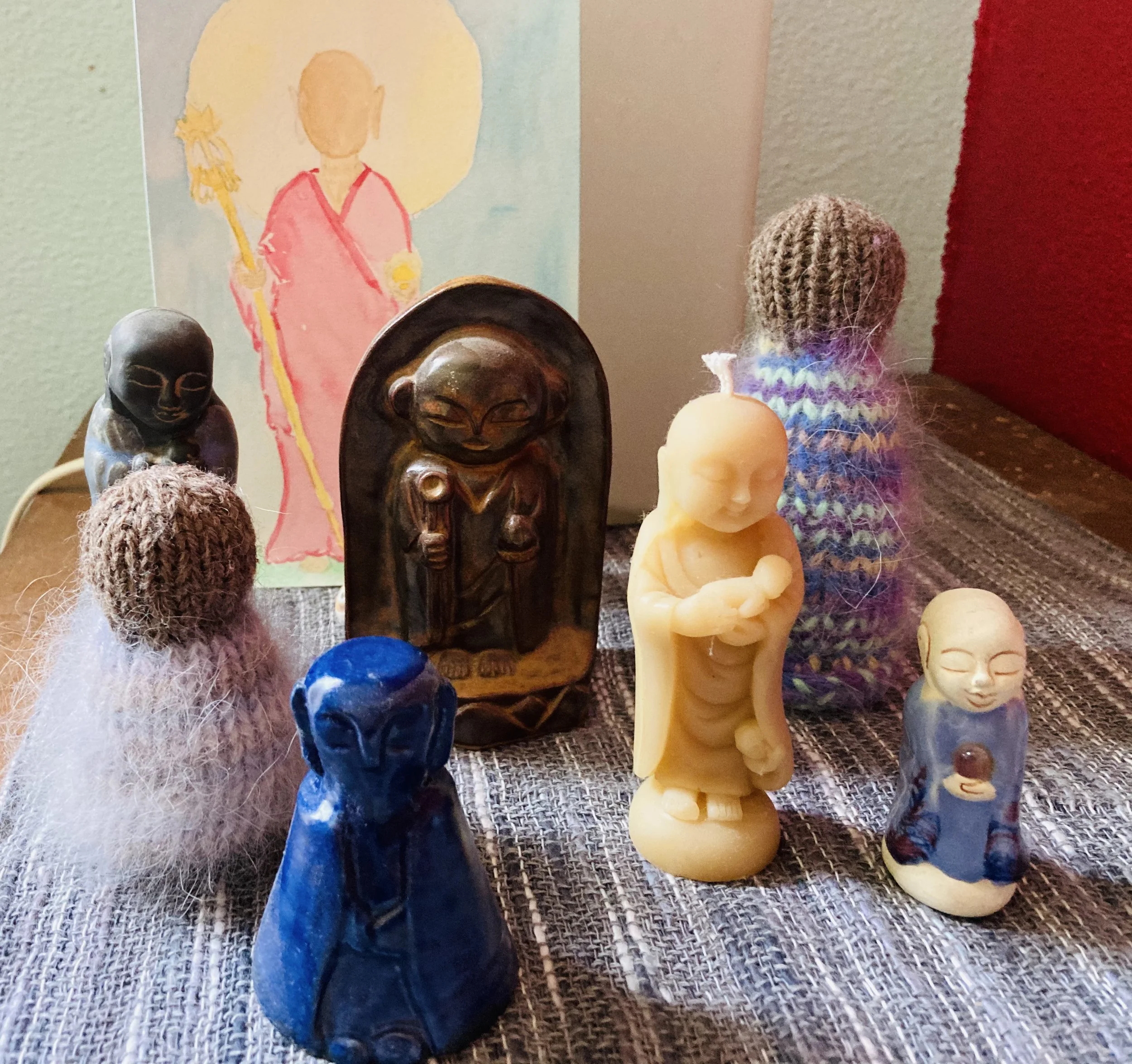 Collection of carved and painted figurines including a blue pair, a wooden person, a person with a hat, and various small figurines, with paintings on the wall behind.