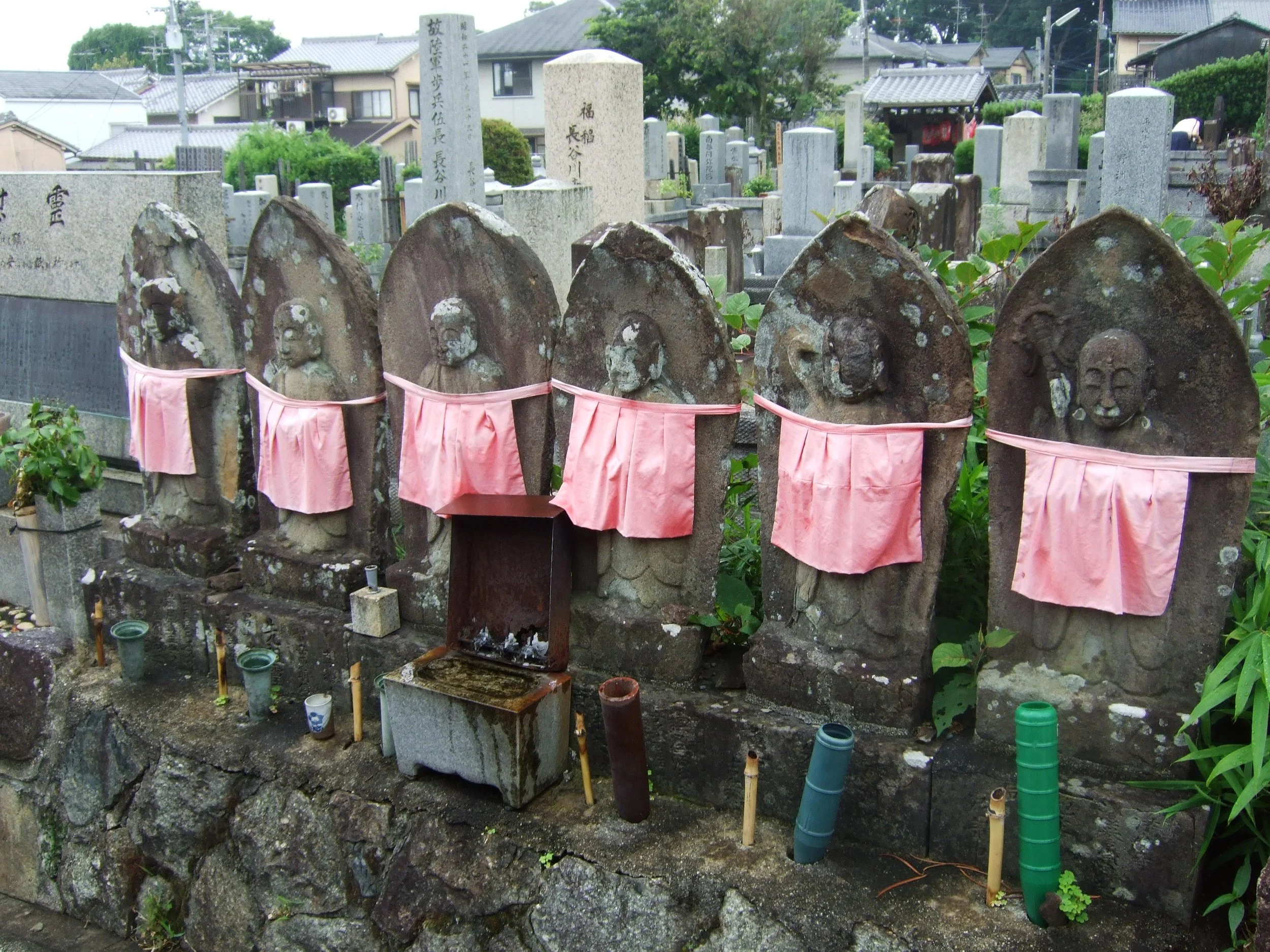 A Japanese graveyard with carved stone Jizo statues wrapped in pink cloth, surrounded by various stone and concrete headstones, some with Japanese inscriptions, and some are weathered and moss-covered, with small plants and colorful pipes nearby.