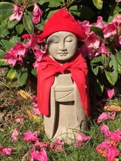 Stone garden sculpture of a peaceful, smiling child with closed eyes, wearing a red hat and red scarf, surrounded by pink flowers.