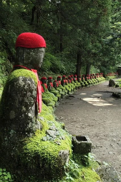 Row of moss-covered stone statues with red caps and red neckerchiefs along a forest path.