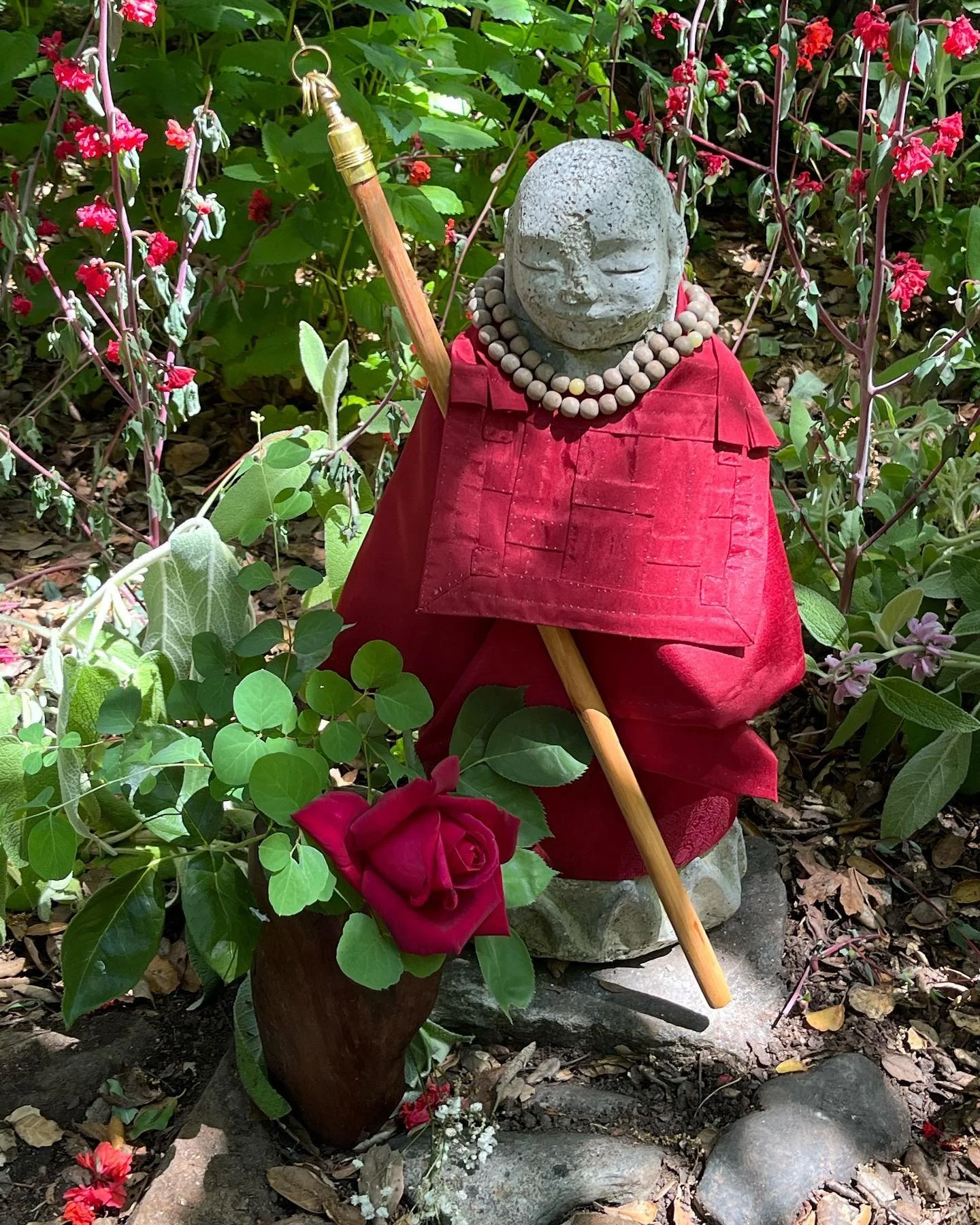 A small stone statue of a smiling monk or child with a beaded necklace and a red robe, holding a staff, surrounded by flowers and green plants in a garden.