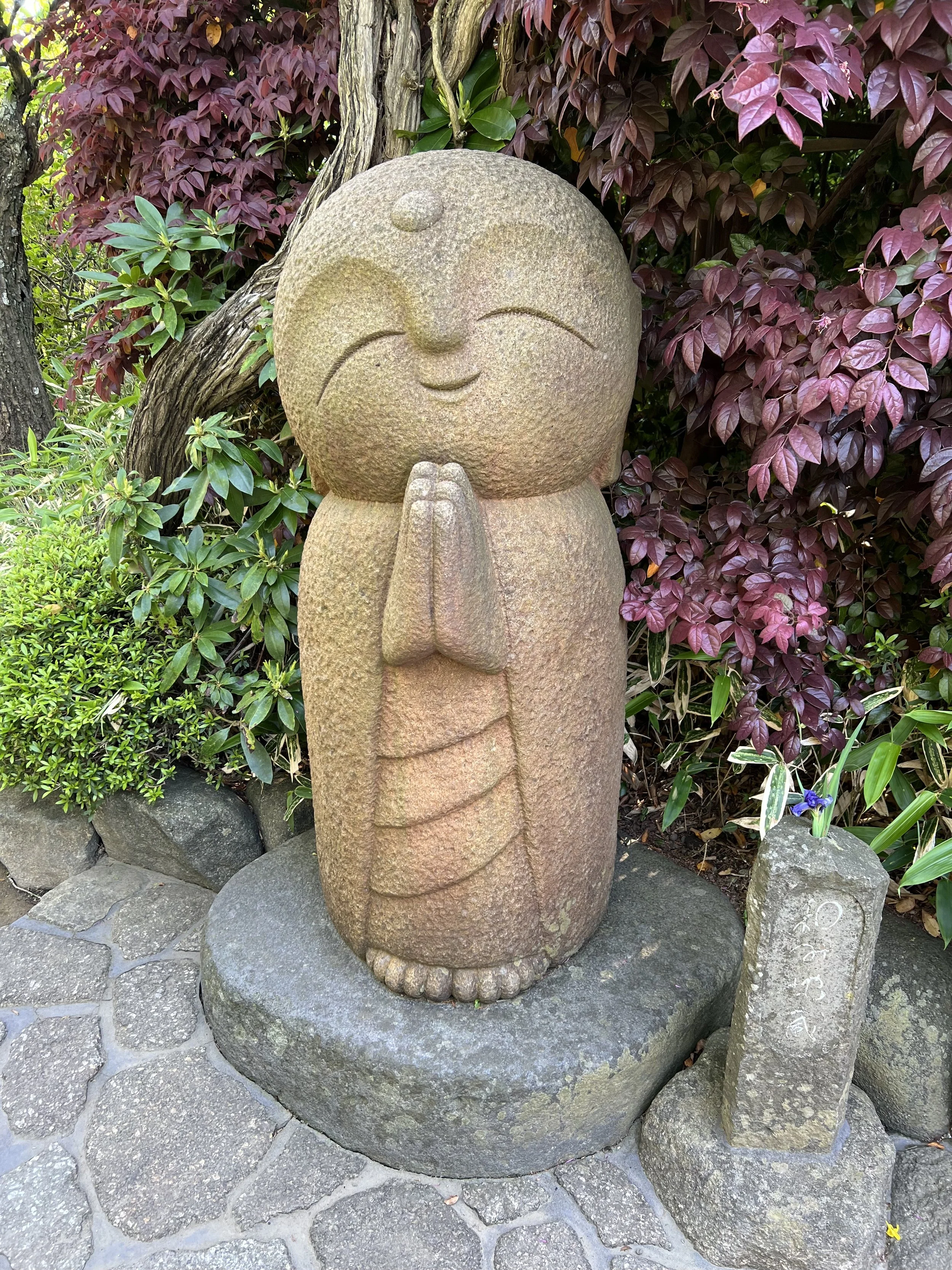 A stone sculpture of a smiling face with closed eyes, hands pressed together in a prayer position, and a rounded head, set against a background of green and purple foliage.