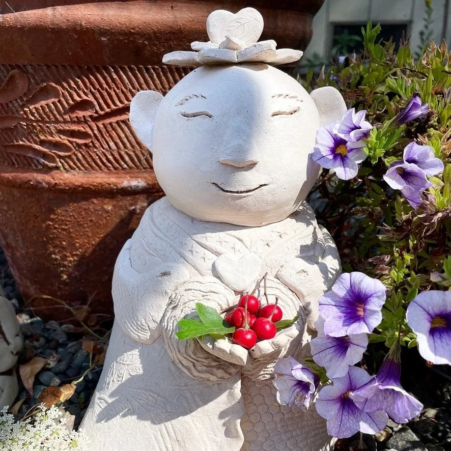 A decorative sculpture of a smiling Buddha-like figure made of white material, holding a small bowl with red cherries and green leaves, surrounded by purple and white flowers and a large terra cotta pot in the background.