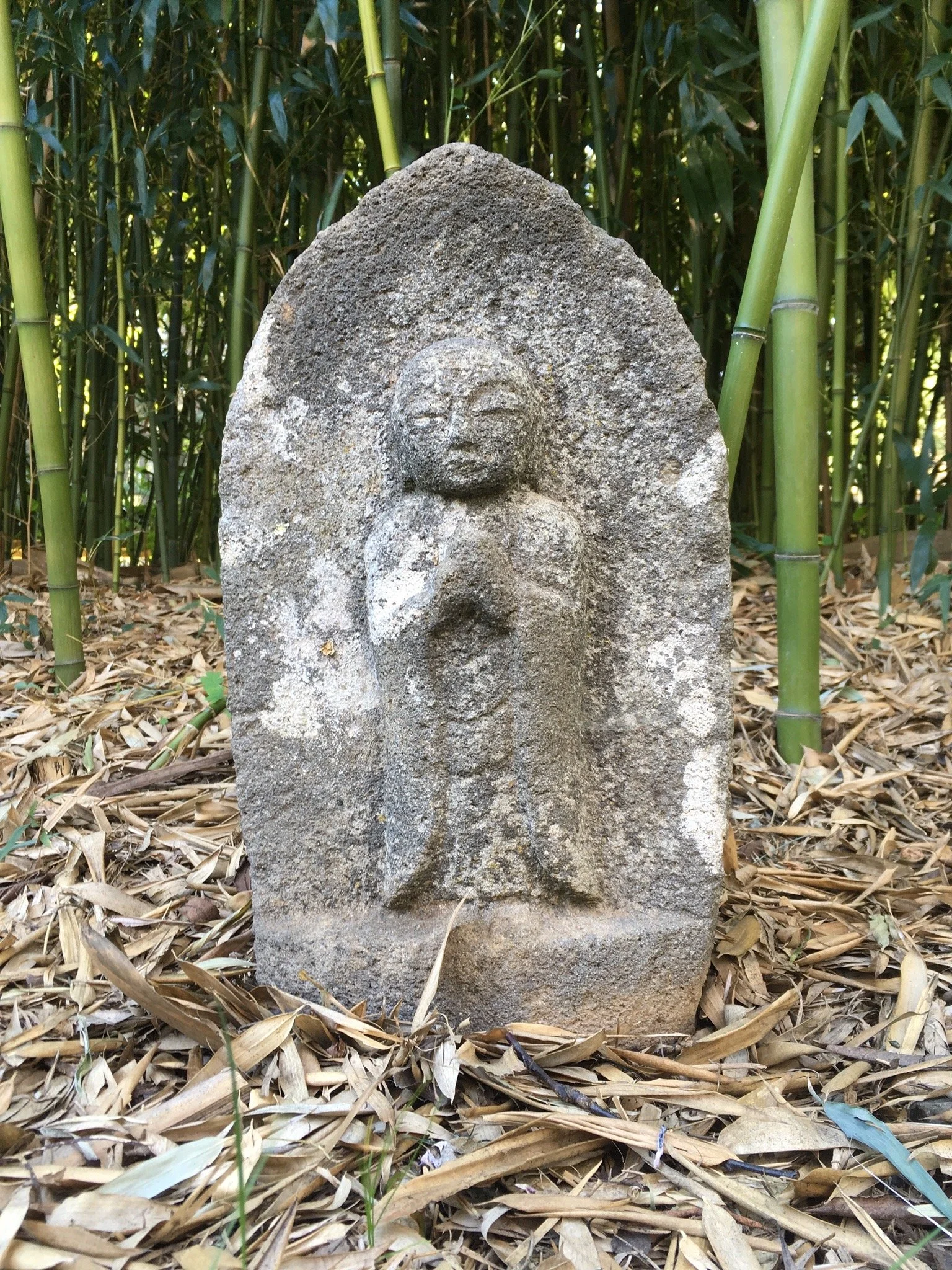 Stone Buddhist statue standing among bamboo stalks and dry leaves on the ground.