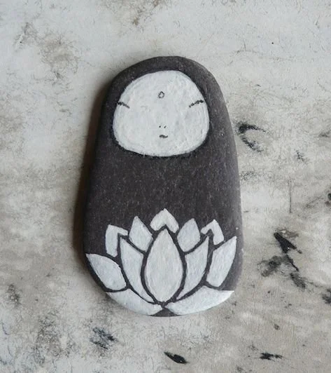 Painted rock featuring a minimalist cartoon face of a girl with closed eyes and a neutral expression, with a lotus flower design at the bottom, on a light textured background.