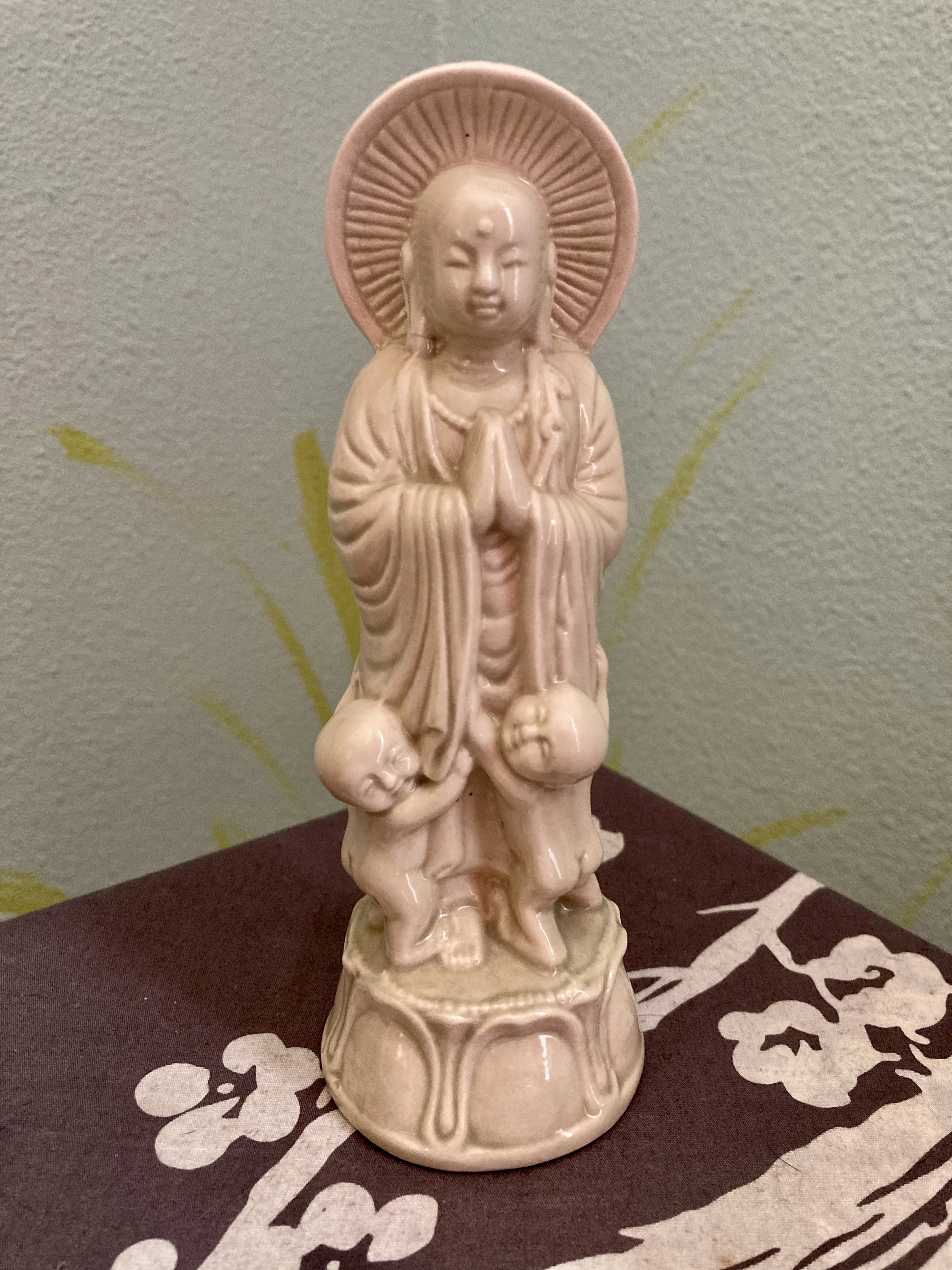 A beige ceramic figurine of a praying monk or deity with a halo behind its head, standing on a carved base, placed on a black and white cloth with floral patterns, against a green wall with faint yellow lines.