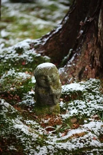 Stone garden statue surrounded by snow and greenery, near a tree trunk.