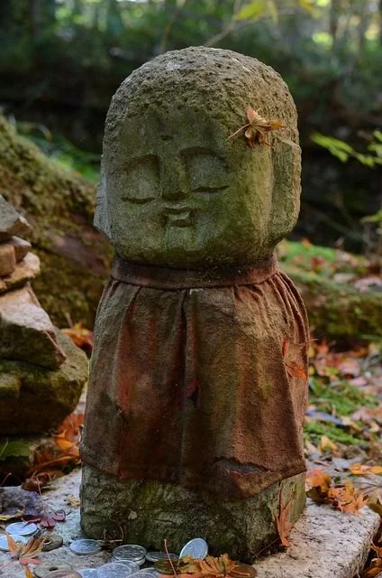 A moss-covered stone statue of a smiling face with closed eyes, wearing a cloth around its neck, with fallen leaves and coins at its base in an outdoor setting.